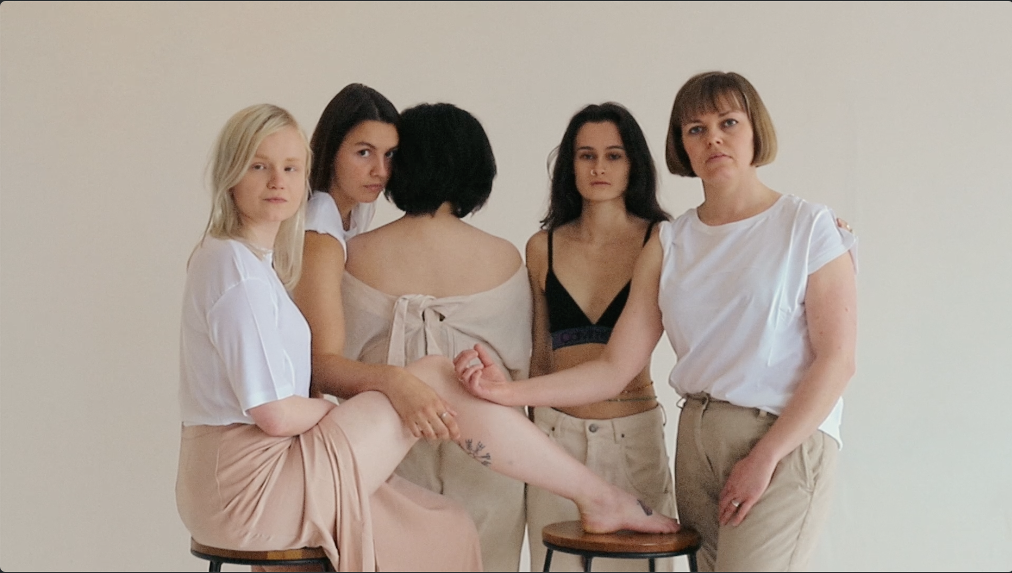 Five women are standing and sitting against a plain white wall. They are dressed in casual clothing, with some wearing white tops and beige pants or skirts. One woman is seated on a stool with her leg raised, and they appear to be posing for a photo with serious expressions.