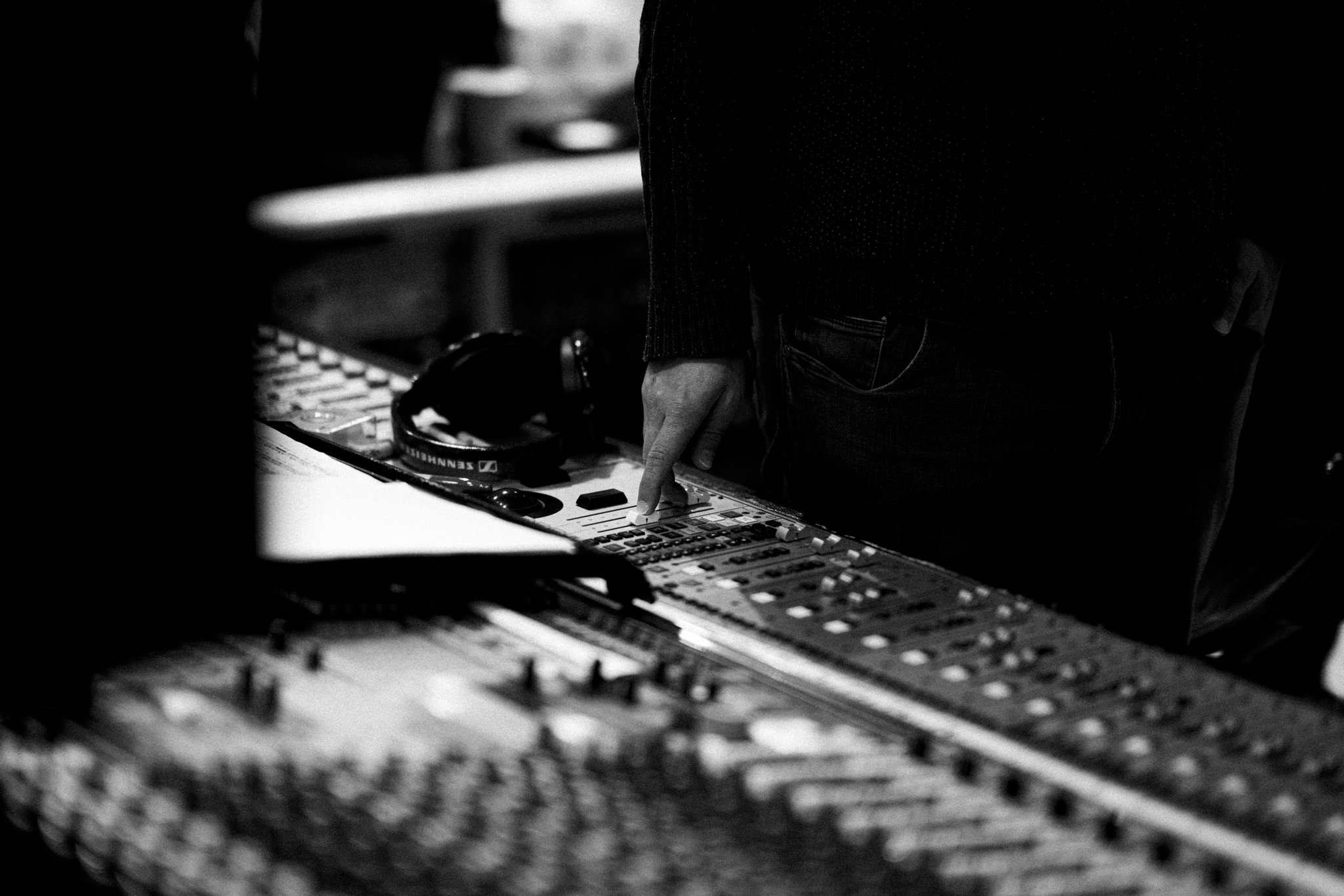A person is standing at a table or desk with electronic equipment, including a mixing console or DJ controller, in a dimly lit room. The person is pointing at the equipment with their right hand.