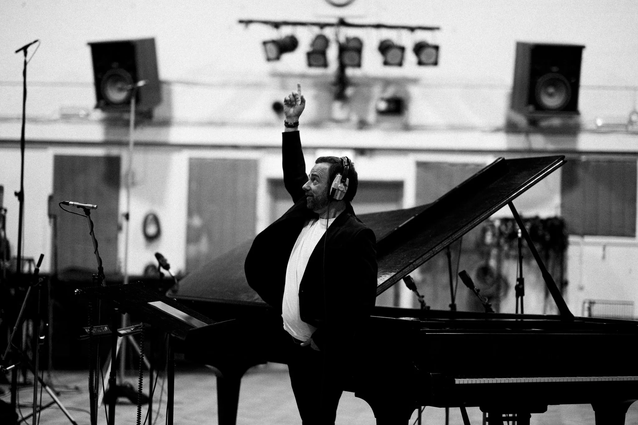 A man wearing headphones playing a grand piano at a recording studio or rehearsal space, with microphones and speakers visible in the background.