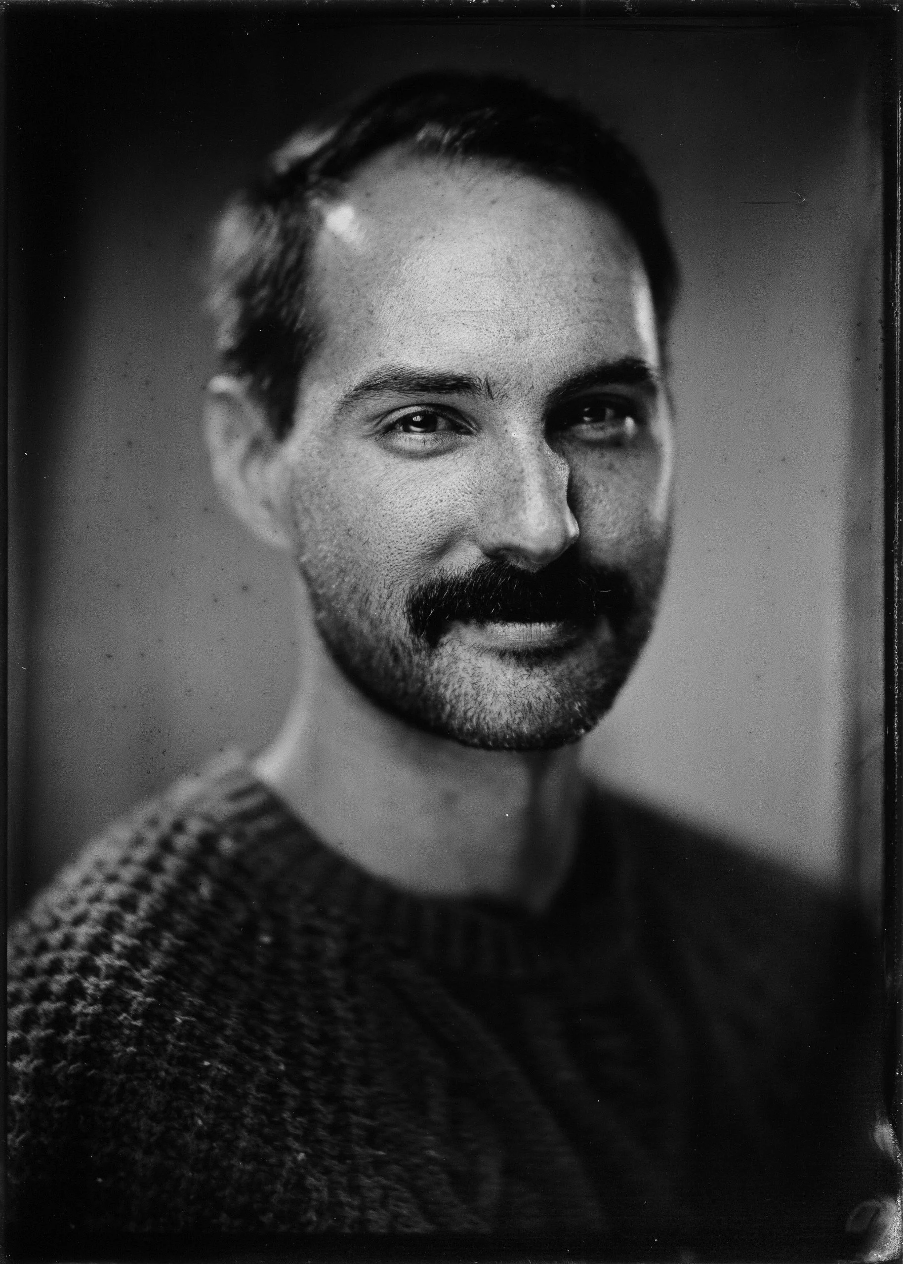 Black and white close-up portrait of a man with dark hair and a beard, smiling slightly, wearing a textured sweater.