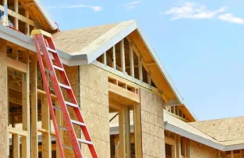 Construction site of a house with wooden framing and a red ladder leaning against the structure, partially finished roof, and blue sky in the background.