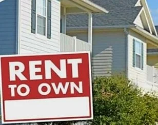 A red and white sign reading 'RENT TO OWN' placed in front of a house with light blue siding and white trim.