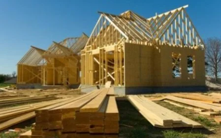 Wooden house under construction with framing and roof trusses installed, construction materials scattered on the ground.