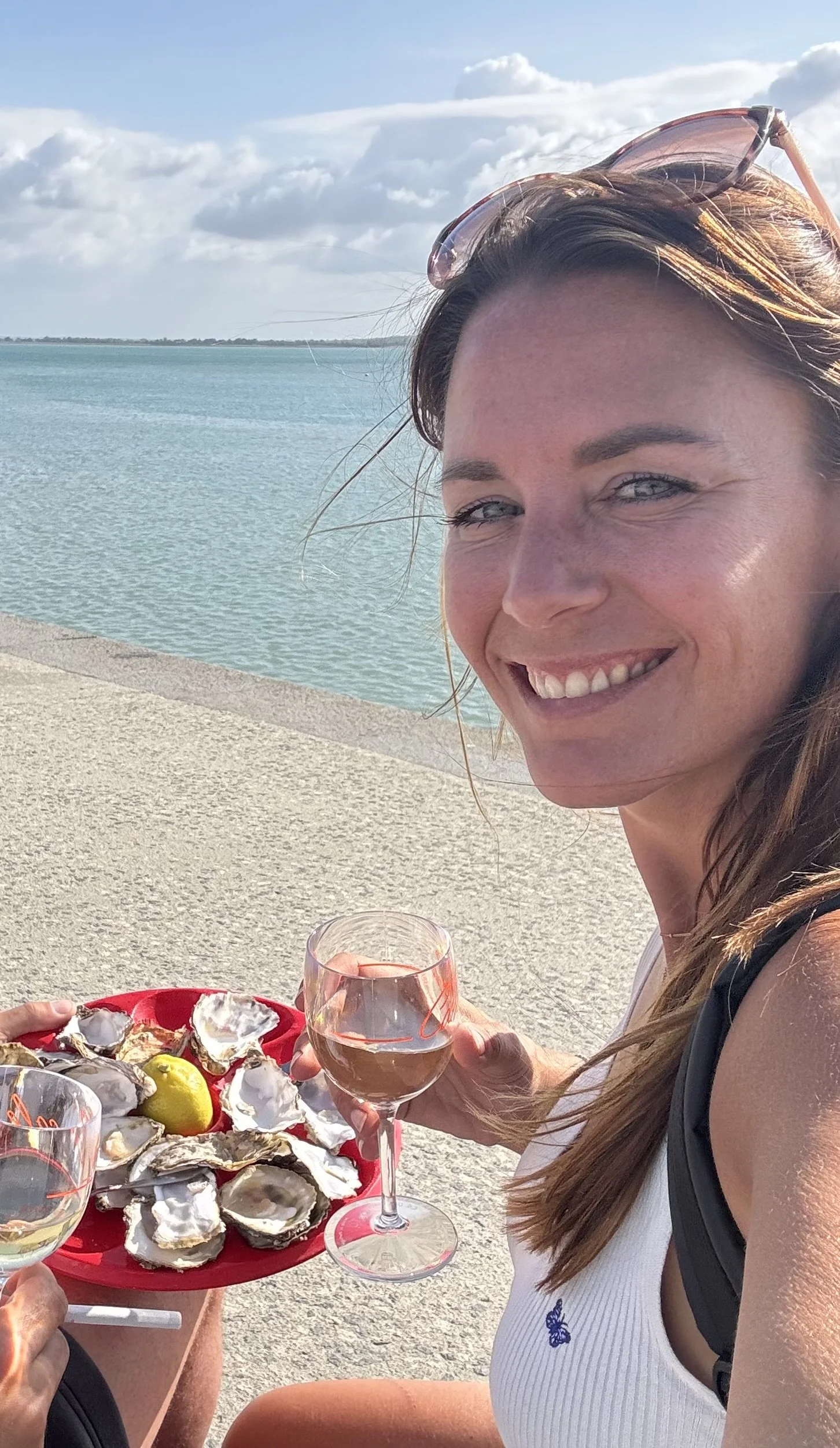 Vrouw (Mieke) met lang bruin haar glimlachend bij zee, drinkend uit een wijnglas, met een schaal oesters en een citroen, op een terras.