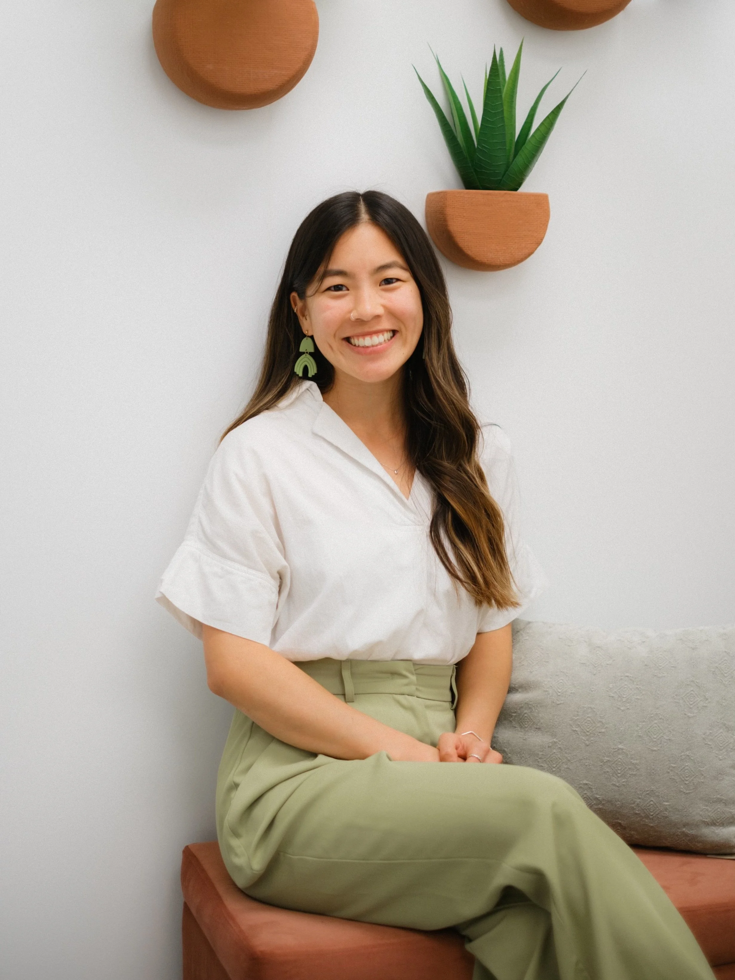 A young woman with long brown hair, smiling, sitting on a pink bench against a plain white wall with wall planters containing a green plant and terracotta semi-circular wall decorations.