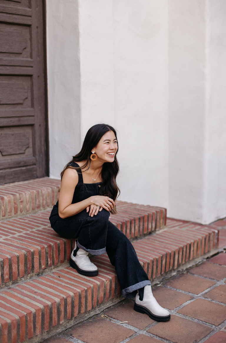 A woman with long dark hair, wearing a black sleeveless jumpsuit, white boots, and gold hoop earrings, sitting on brick steps and smiling outdoors.