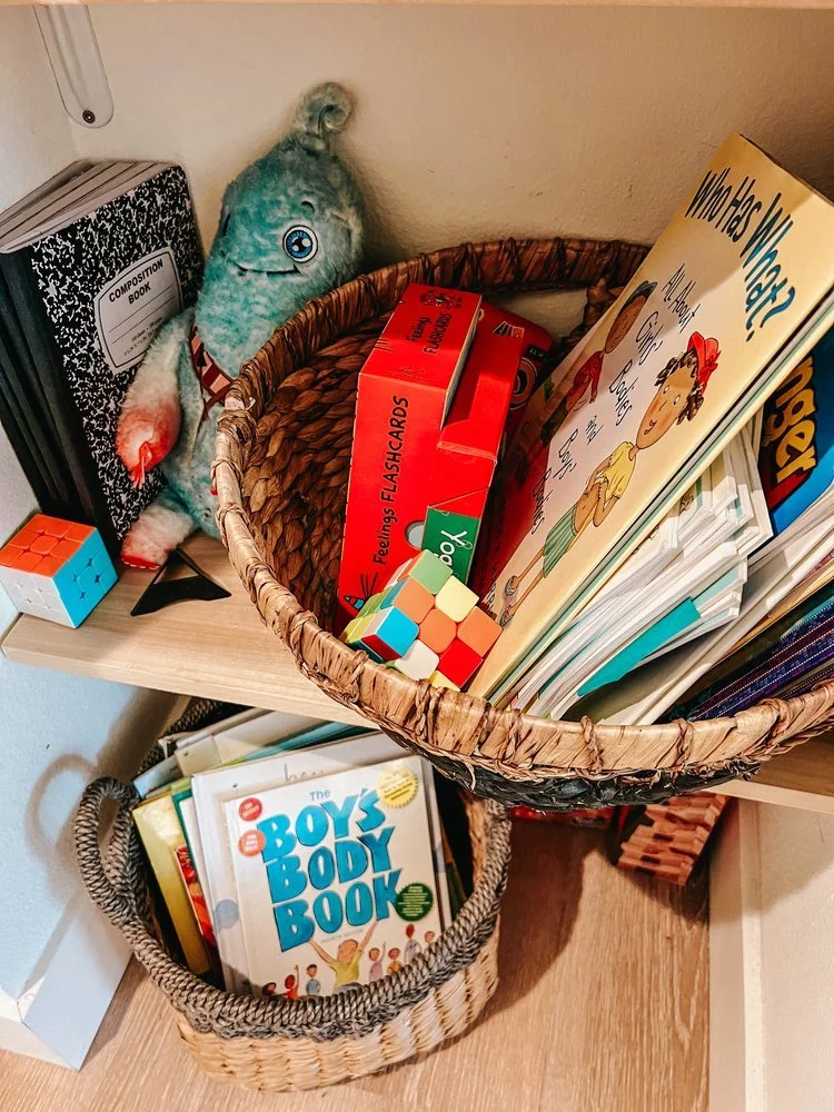 Children's books and toys on a wooden shelf, including a plush doll, a Rubik's Cube, Flashcards, and books about health and body awareness.