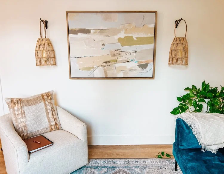 Living room corner with beige armchair, plaid pillow, notebook, and pen, a blue velvet sofa with a white fringe blanket, green plant, abstract wall art, and wall-mounted wicker baskets.