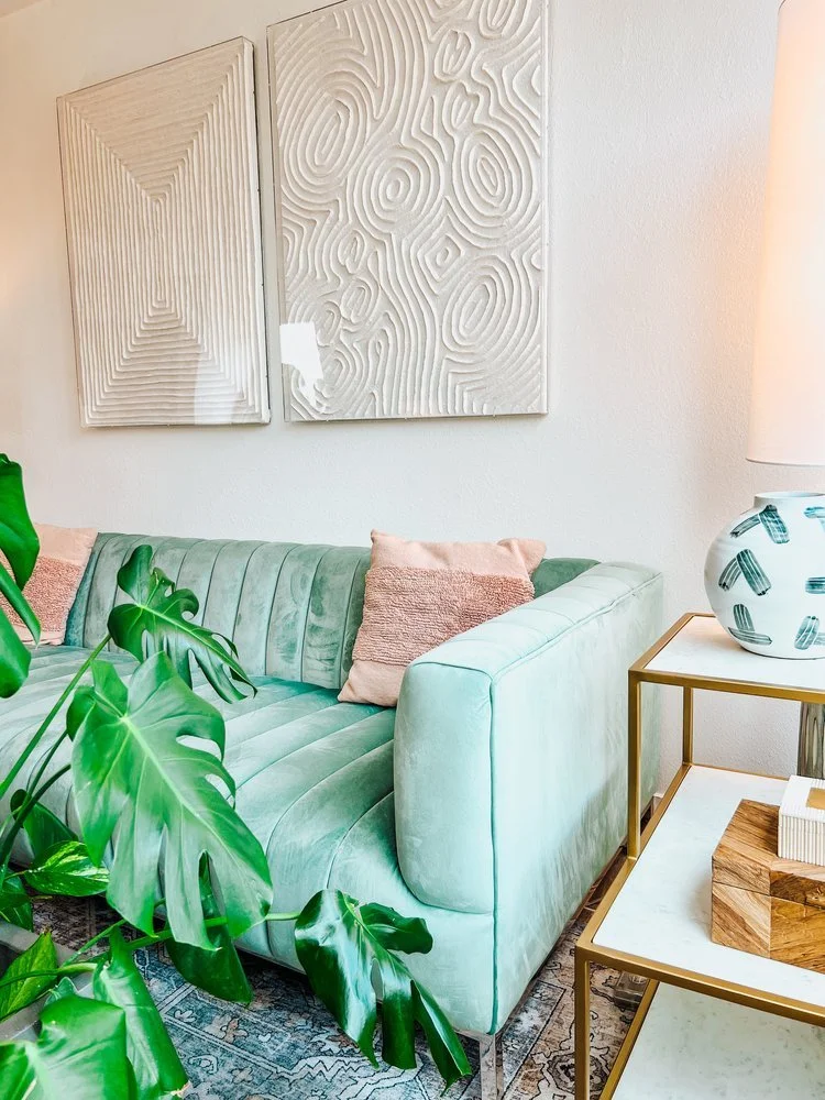 Living room with a mint green velvet sofa, decorative pillows, a side table with a large white and blue patterned vase, and abstract wall art, along with a green plant in the foreground.