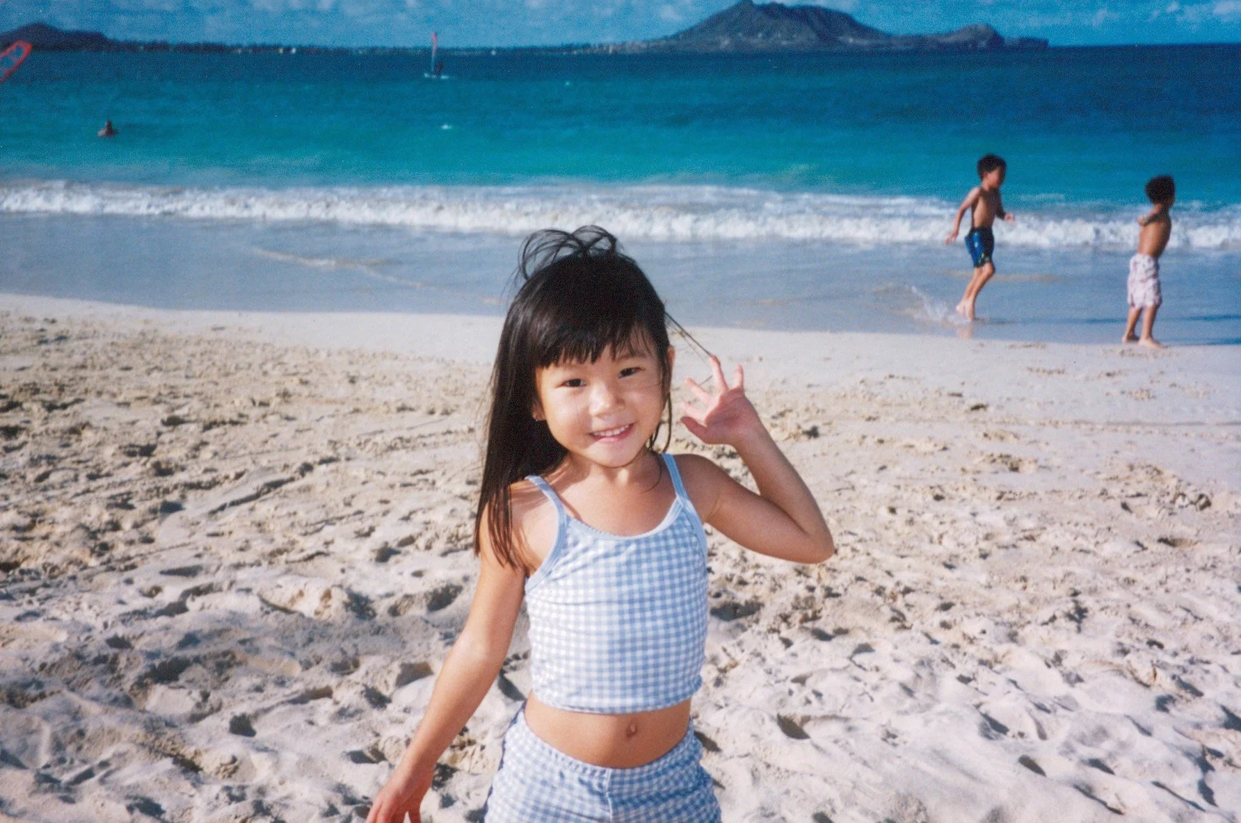 A young girl with long dark hair, smiling and waving on a sandy beach with the ocean and a mountainous island in the background. Two shirtless boys are playing in the shallow water, and there are sailboats visible in the distance.
