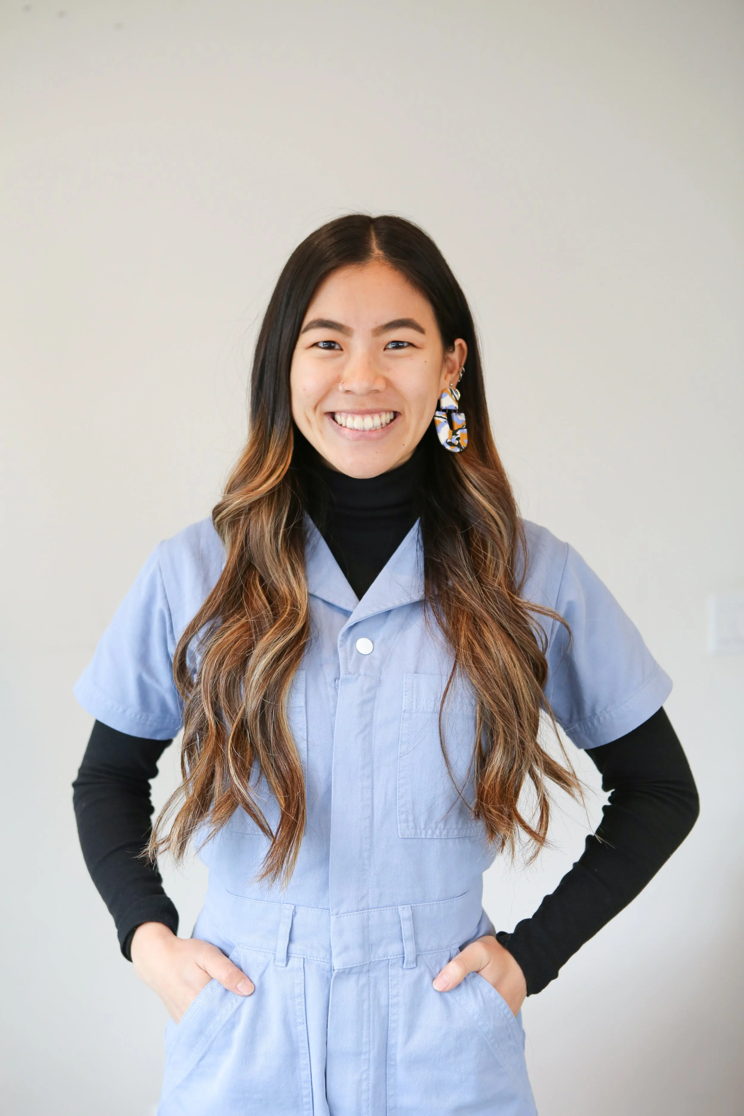 Smiling woman with long wavy hair wearing a light blue medical scrub top, black long sleeve shirt underneath, with hand in her pocket, standing against a plain wall.