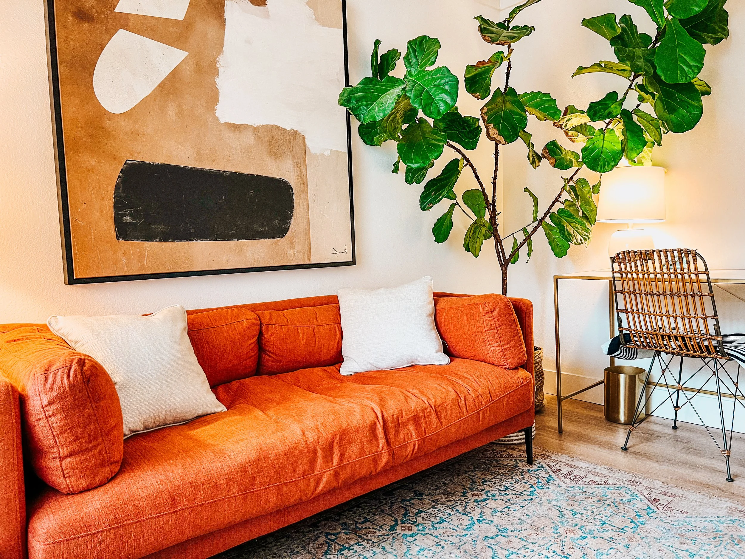 Living room interior with an orange sofa, abstract wall art, a large potted plant, and a desk with a rattan chair.