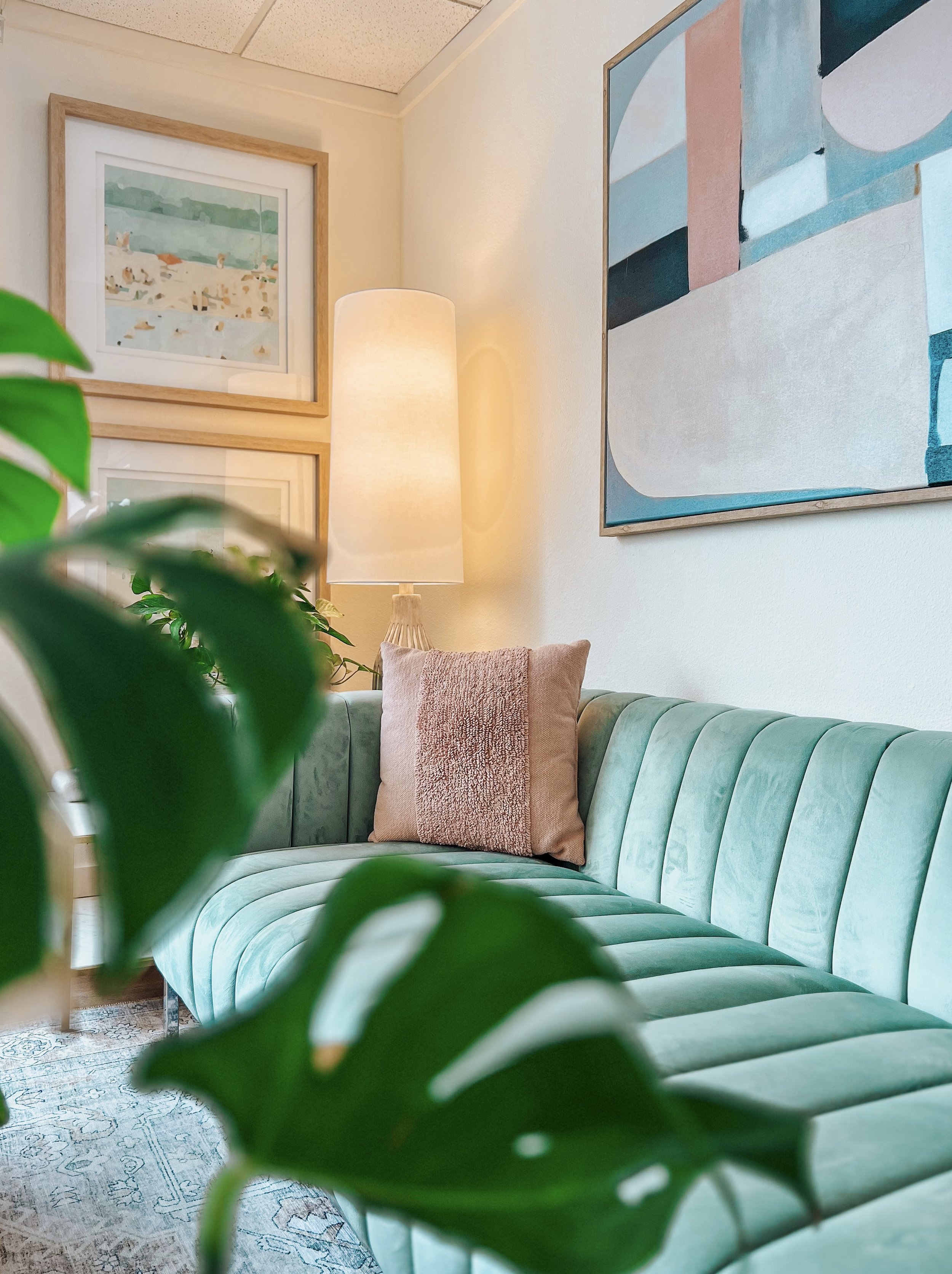 Stylish living room with a teal velvet sofa, beige pillow, abstract wall art, large floor lamp, and plant leaves in the foreground.