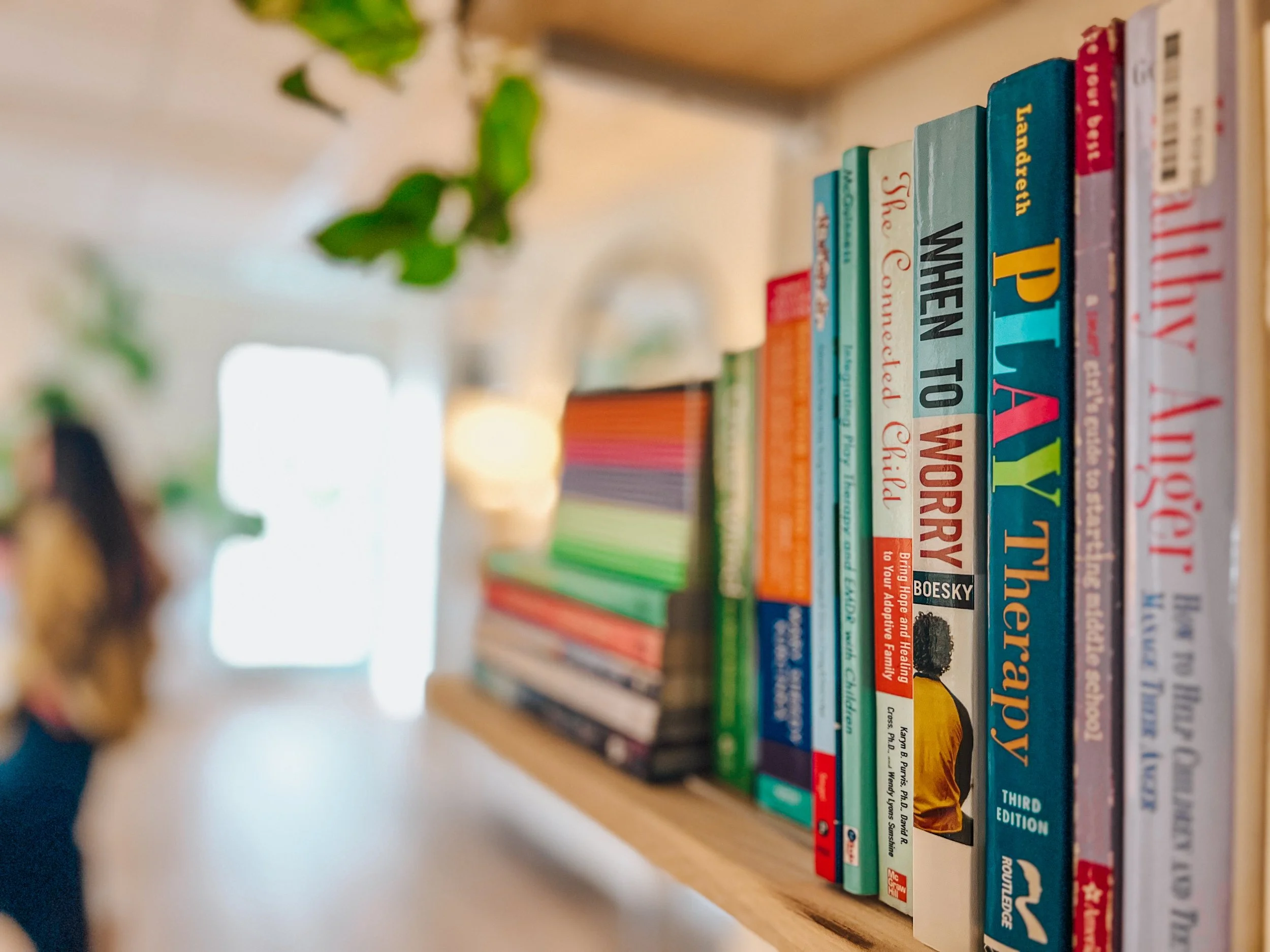 A bookshelf with various colorful books, including titles about psychology and therapy, with a blurred background showing plants and a person.