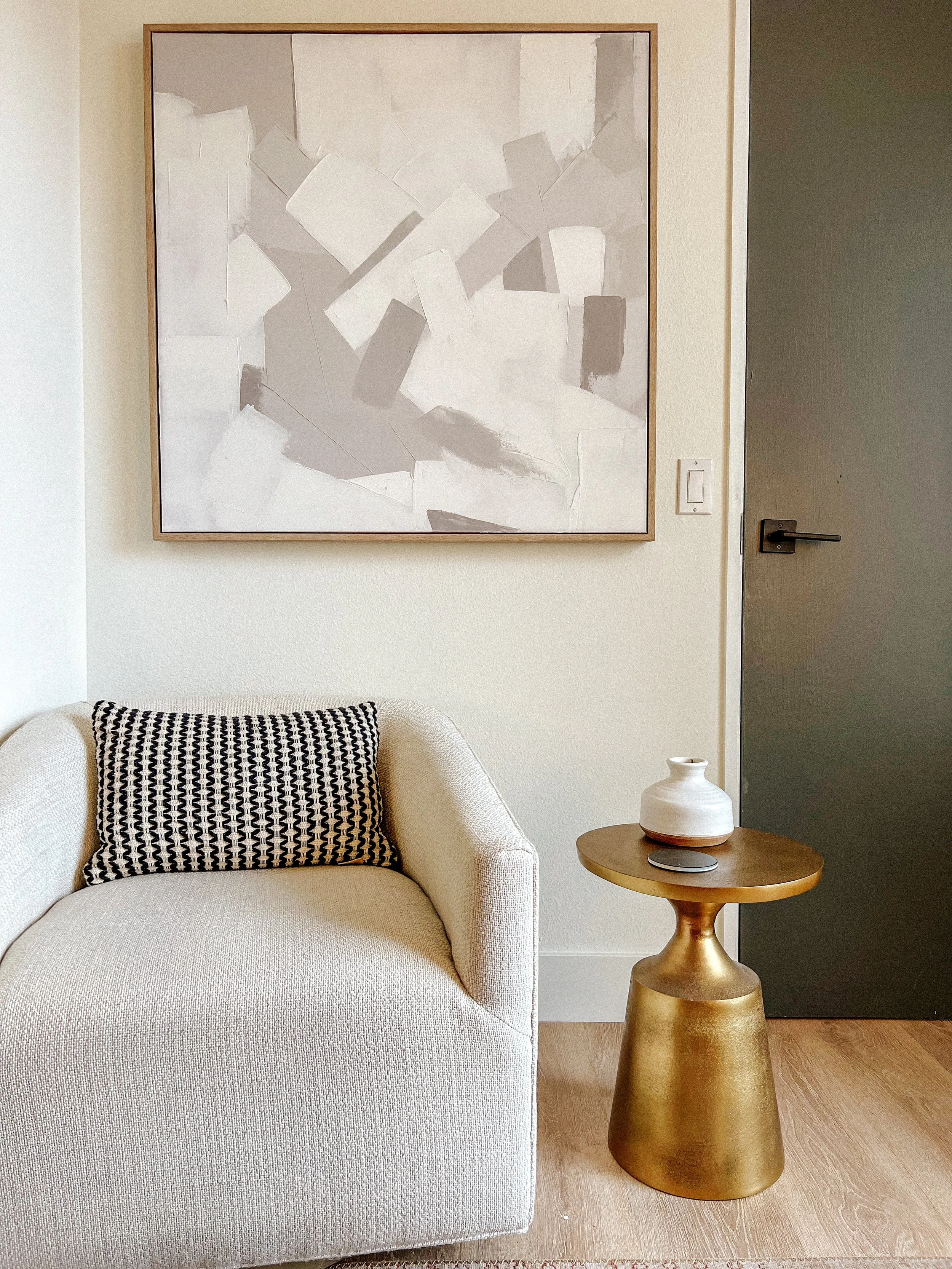 Minimalist living room with a beige armchair, a black and white patterned pillow, a small gold side table holding a vase, and abstract artwork on the wall.