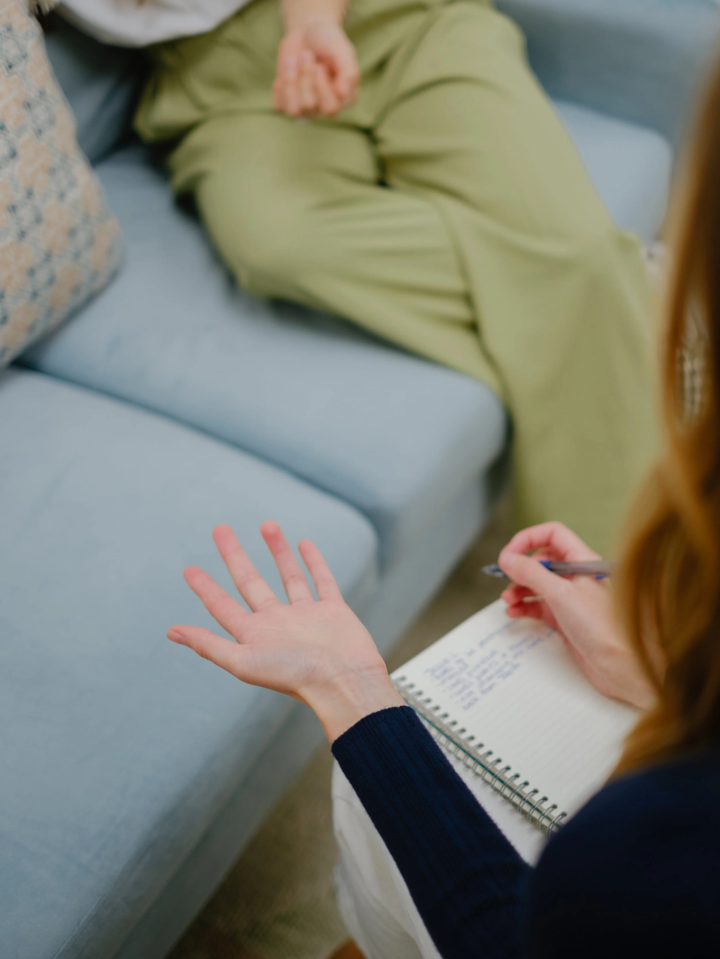 A person with long red hair wearing a navy sweater is writing in a spiral-bound notebook with blue lines. Another person, sitting on a gray couch wearing olive green pants, has their hand extended with the palm facing up, and their other hand is resting on their knee. The scene suggests a casual or work-related setting.