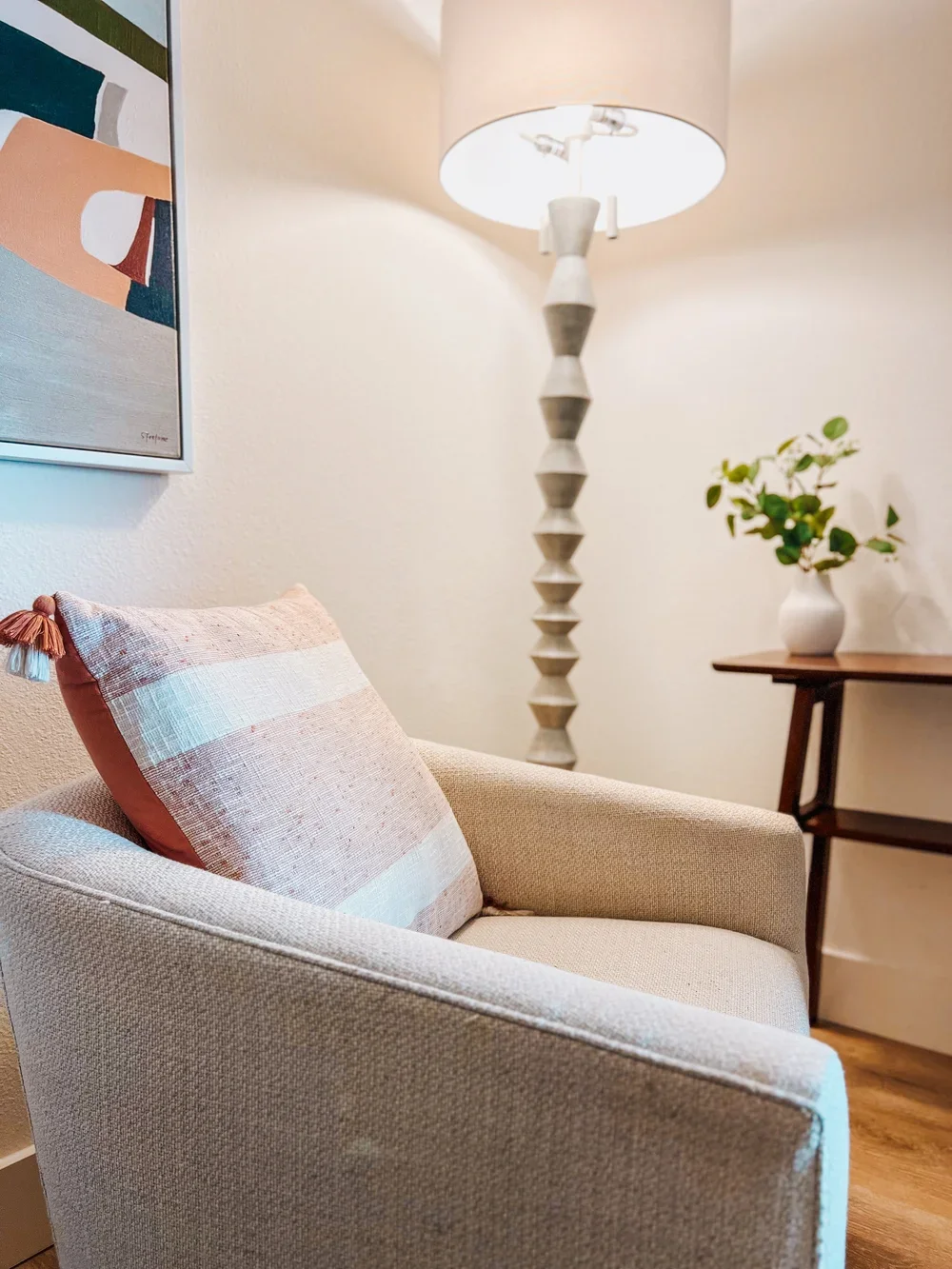 A cozy beige armchair with a pink striped pillow, a wooden side table with a white vase and green plant, a tall modern floor lamp, and colorful abstract artwork on a white wall.