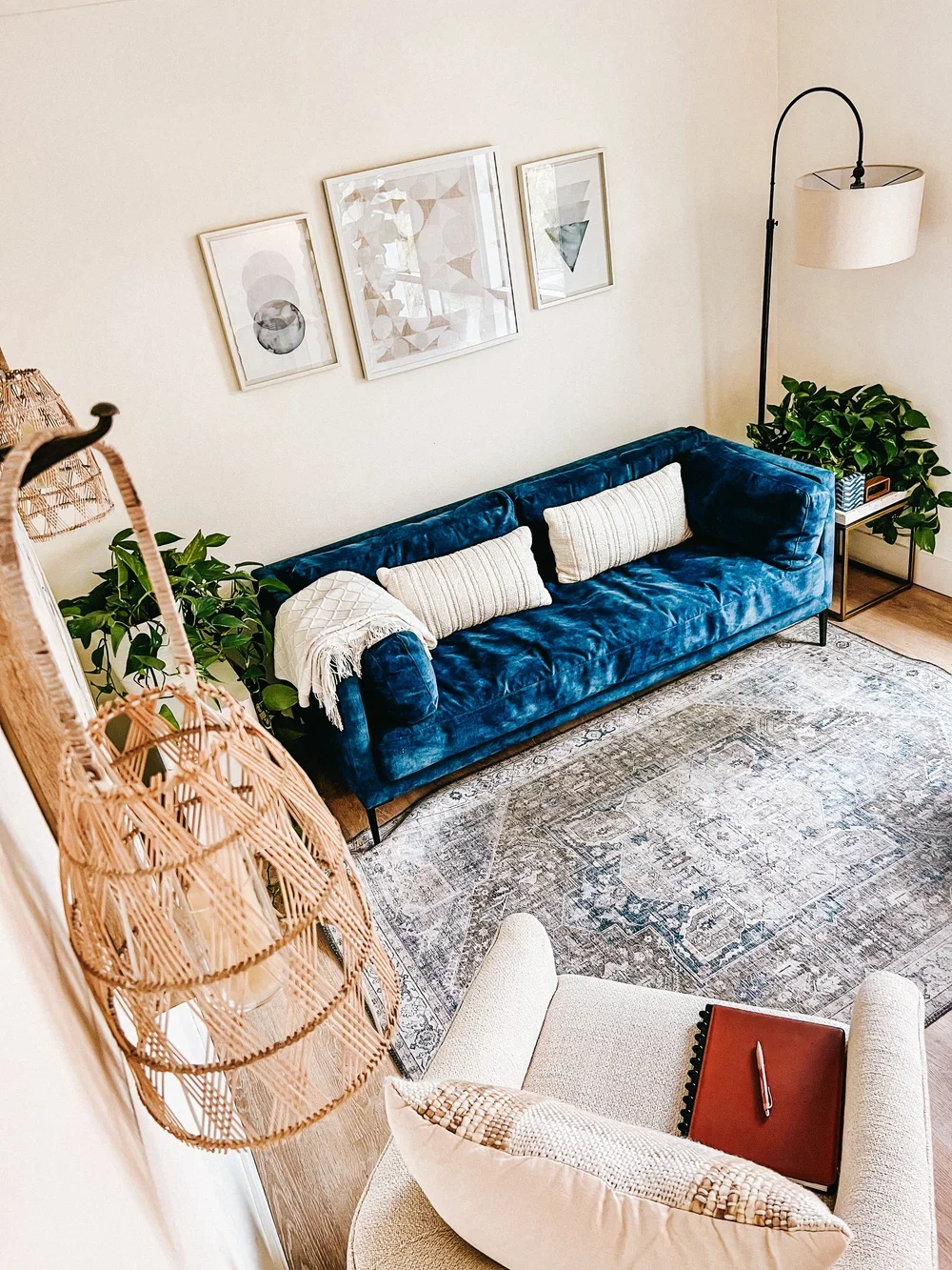 Living room with a blue velvet sofa, beige armchair, patterned area rug, wall art, potted plants, side table, and floor lamp.