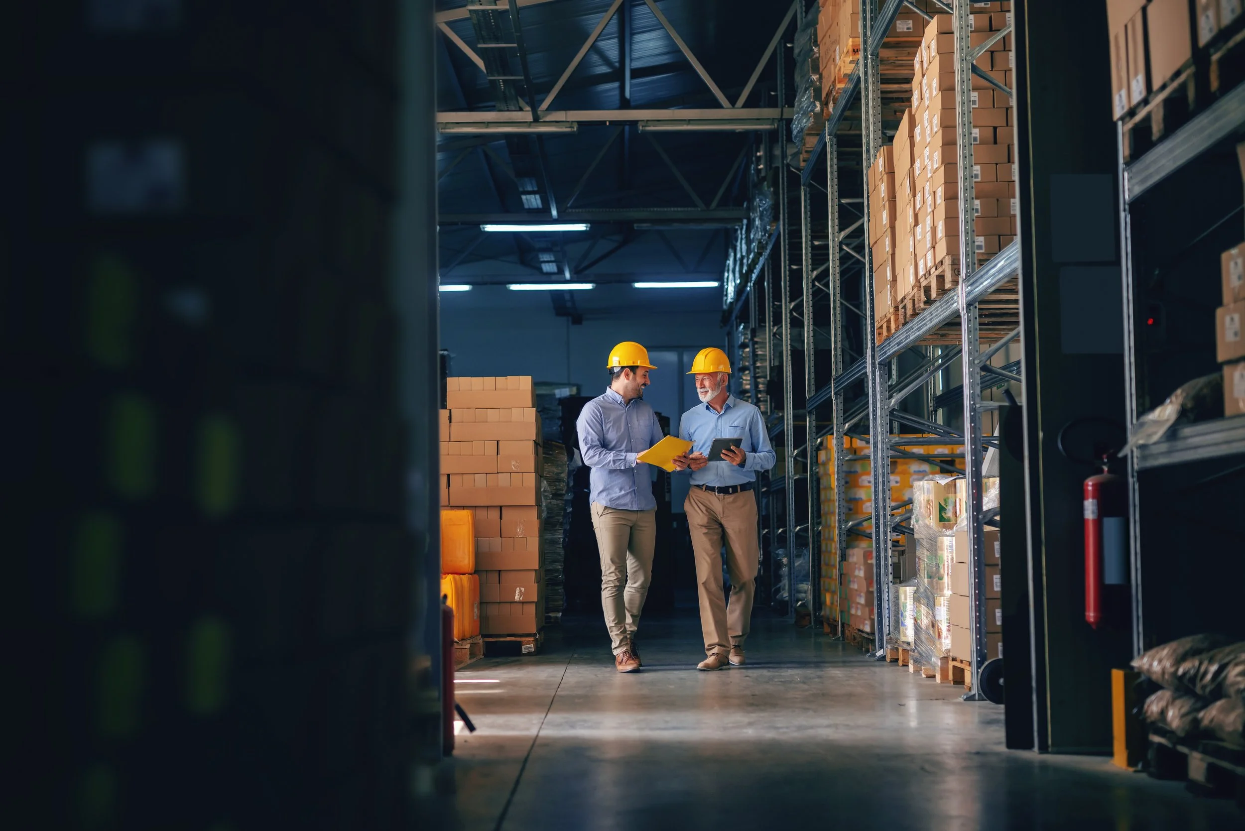 Two warehouse professionals wearing hard hats reviewing documents and a tablet beside inventory shelves.