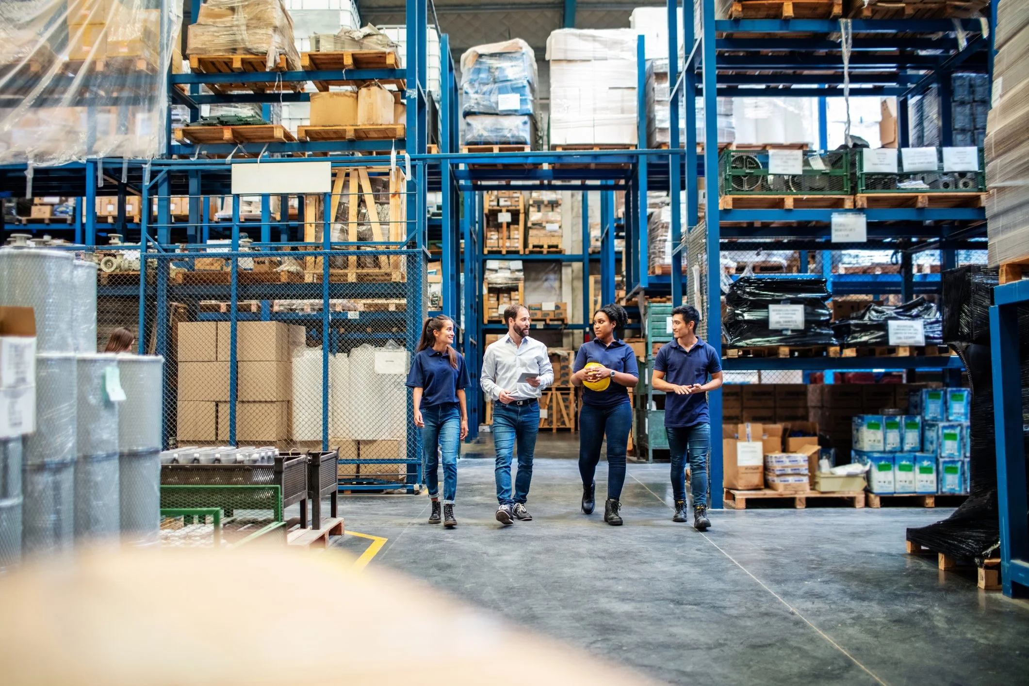 Four business professionals walking through a warehouse with industrial shelving and inventory.