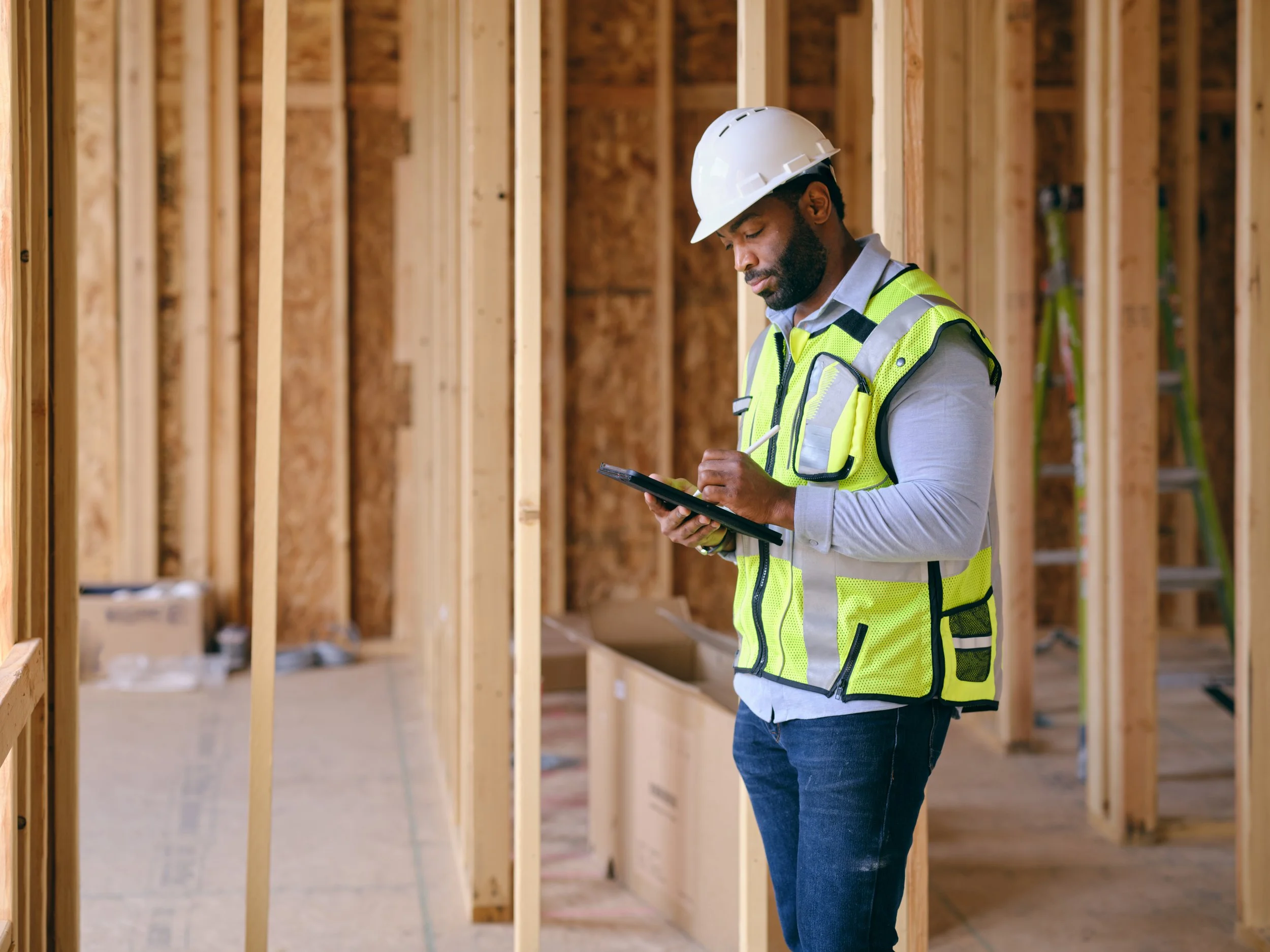 Construction professional wearing a hard hat and safety vest using a tablet inside a wood-framed building.