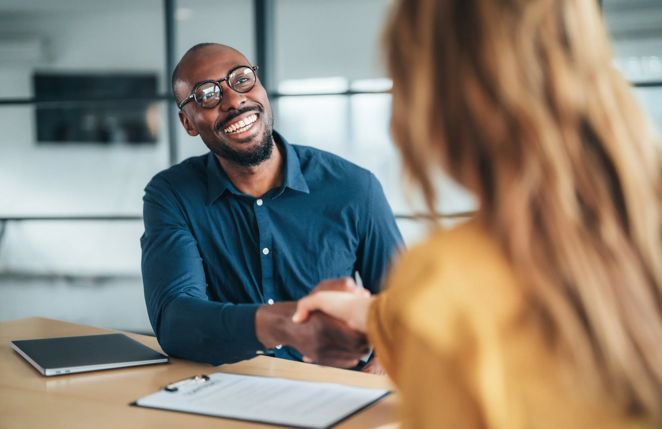 Smiling business professional shaking hands with another person across an office desk.
