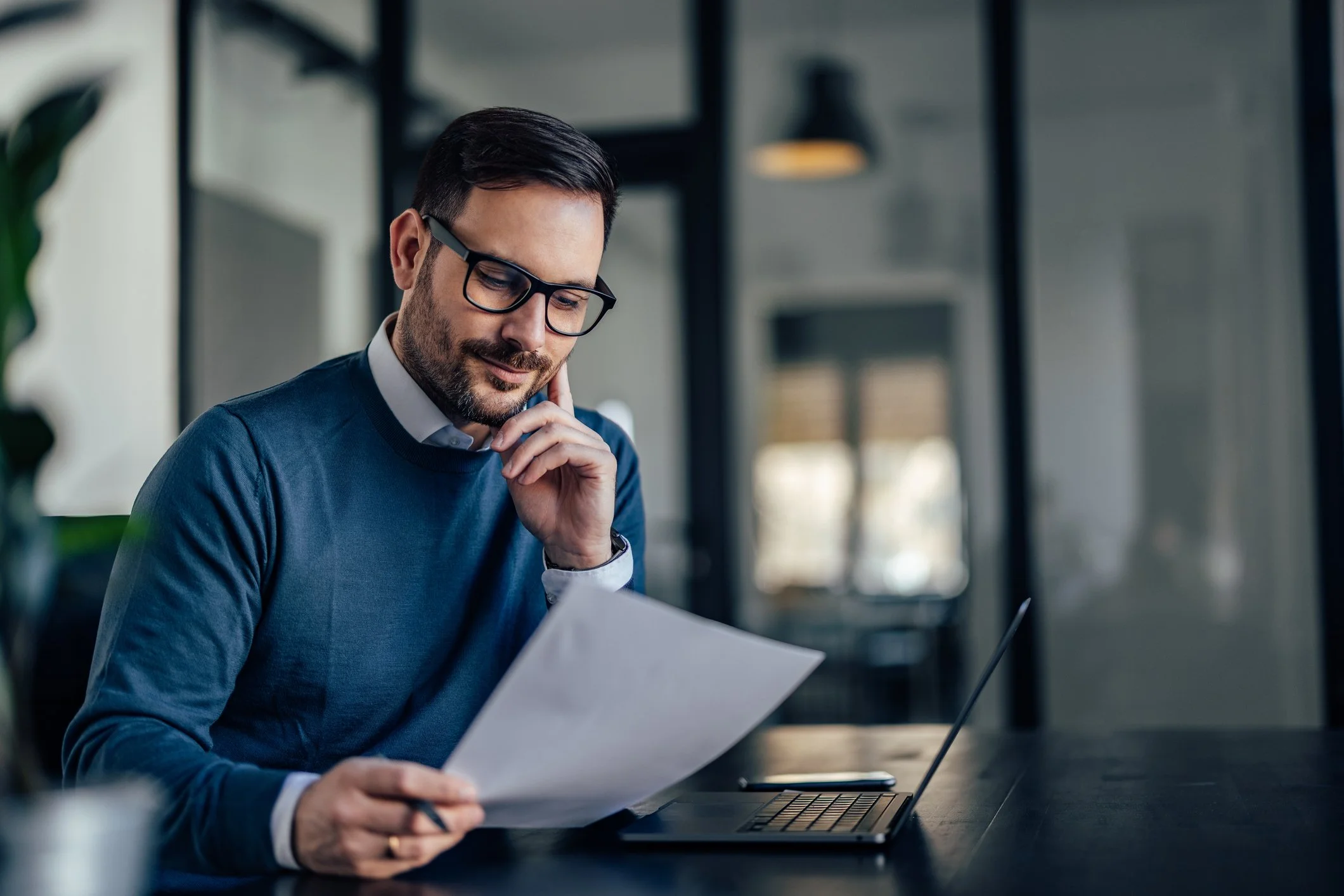 Business professional reviewing financial documents at a desk with a laptop.