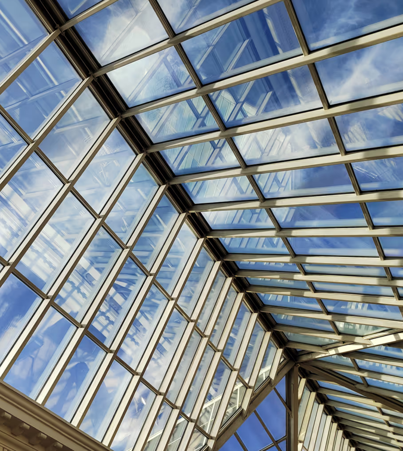 Perspective view of a glass ceiling with a geometric pattern of glass panels and metal supports, showing reflections of the sky and clouds.