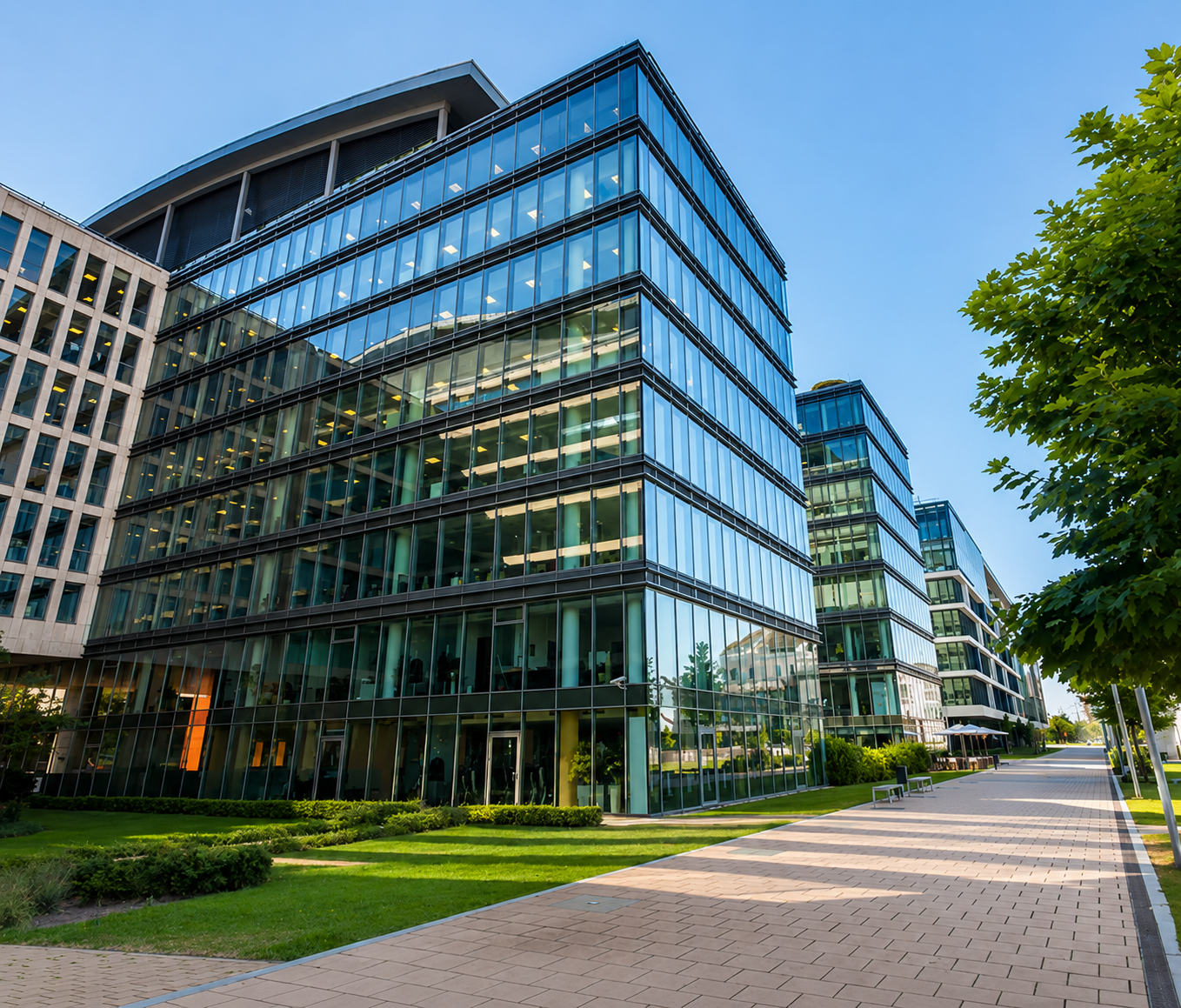 Modern office buildings with glass facades along a sidewalk with benches, trees, and landscaped grass under a clear blue sky.