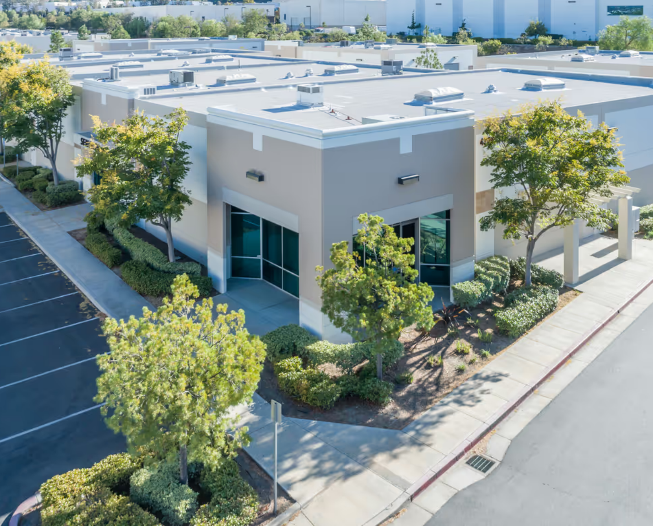 Aerial view of a commercial building with a flat white roof, surrounded by green trees, a parking lot, and sidewalk, with other large white industrial buildings in the background.