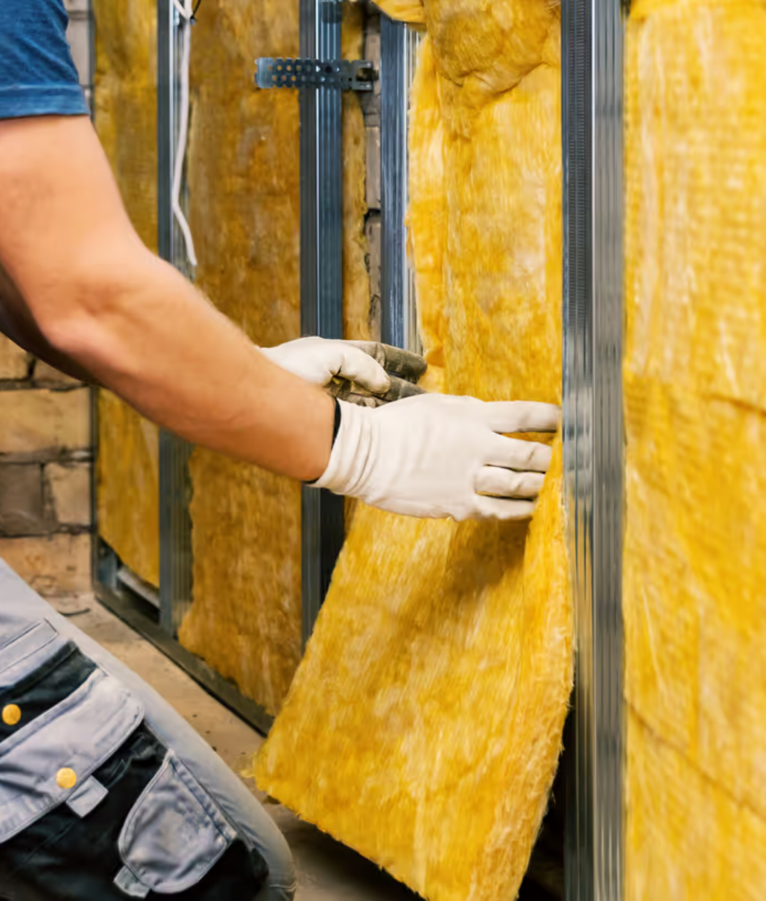A person installing gold-colored insulation foam panels on a wall with metal framing, wearing work gloves and work boots.