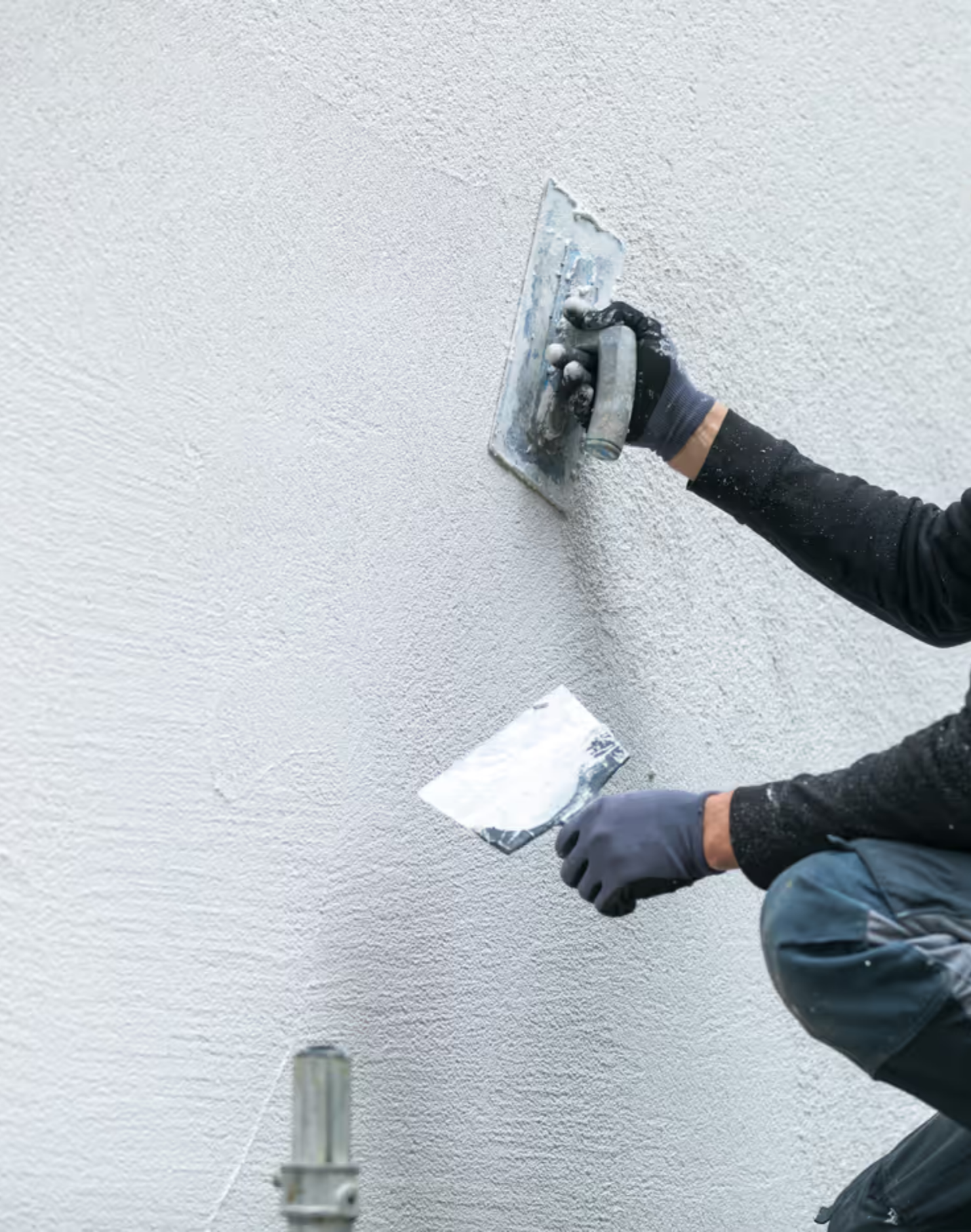 A person is applying textured white paint to a wall with a trowel, wearing gloves and dark clothing.