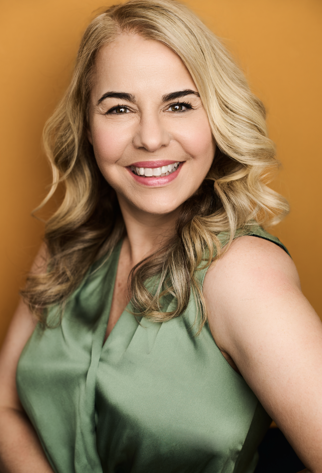 A smiling woman with long, styled blonde hair wearing a sleeveless green top, posing with a yellow background.