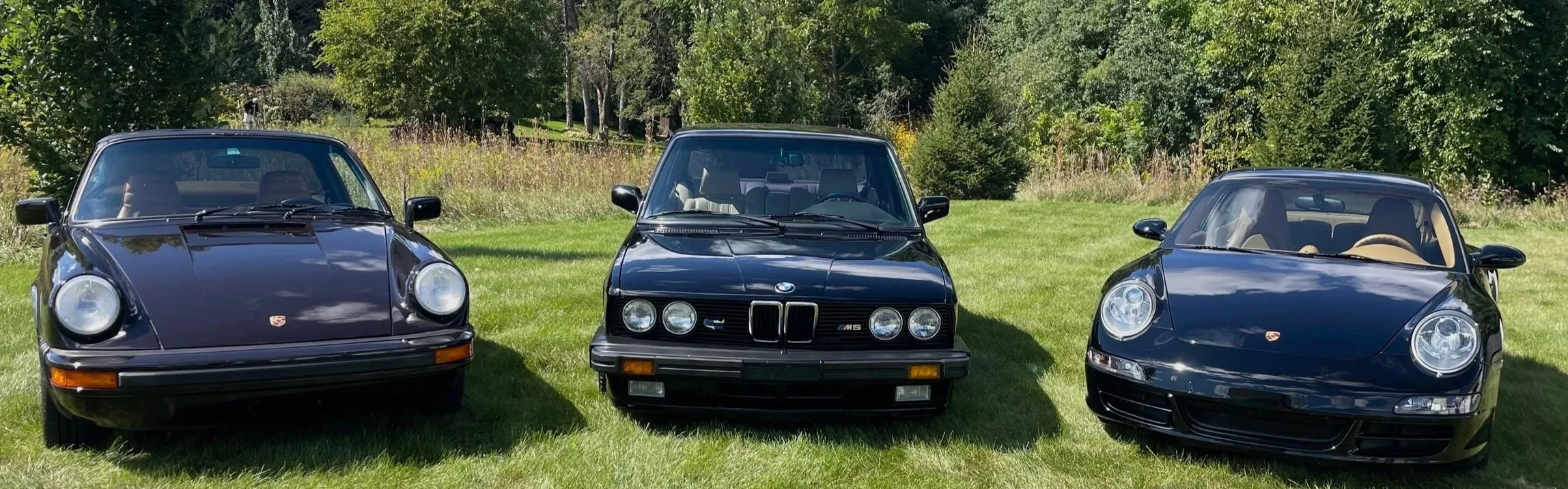 Two vintage Black Porsche 911s and a classic BMW M5  parked on a grassy field with trees in the background, showcasing classic and modern models.