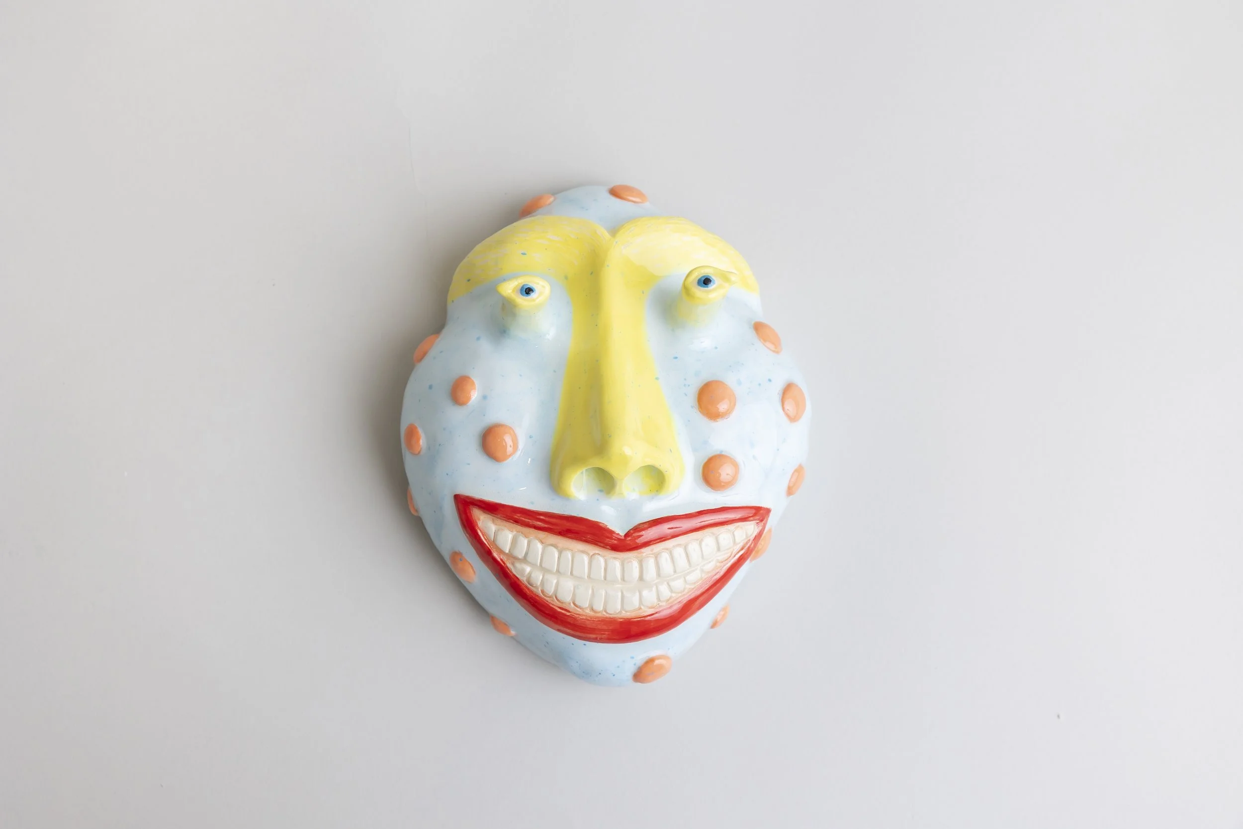Colorful ceramic mask with a smiling face, blue and yellow face, orange spots, blue eyes, red lips, and a white background.