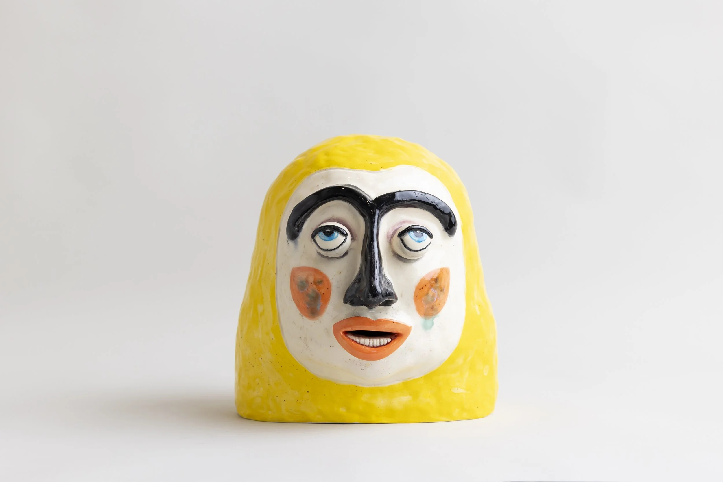 Ceramic figurine of a person's head with a painted face, yellow hair, and exaggerated facial features, set against a plain light gray background.