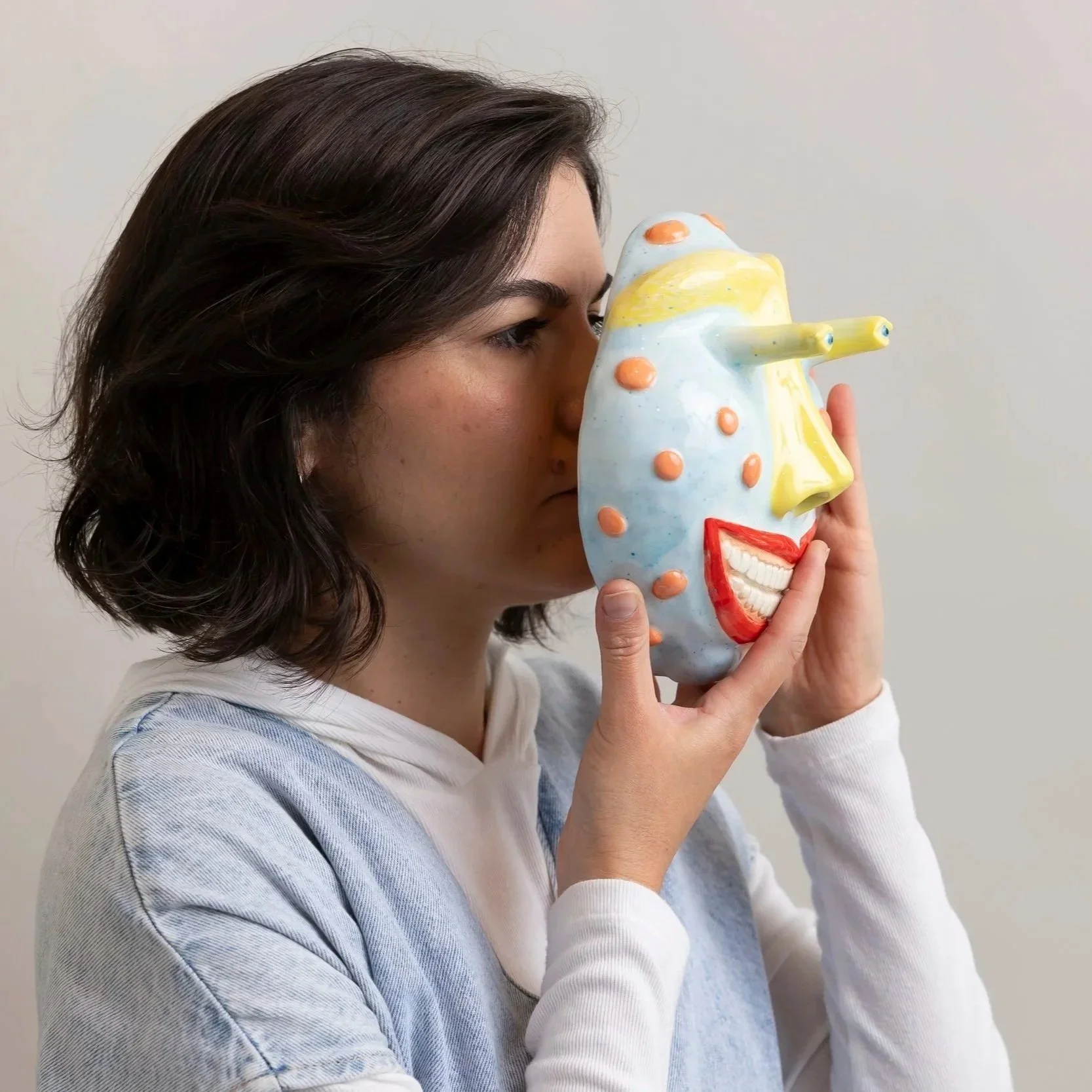 A woman with dark hair holding a colorful ceramic mask up to her face, looking to the side against a plain wall background.