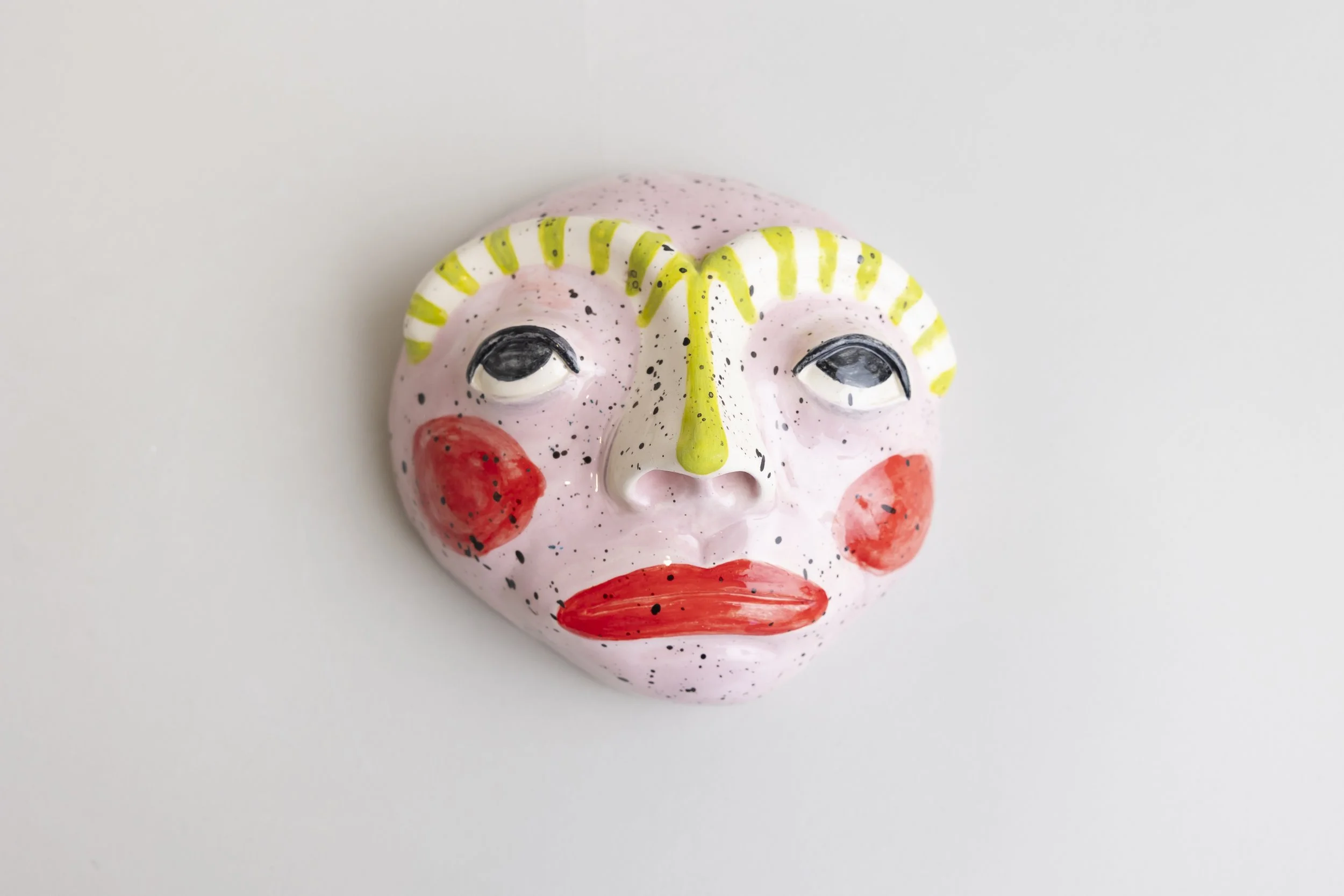 A ceramic mask painted with a pink face, black speckles, red cheeks, red lips, and yellow and white patterns around the eyes, on a white background.