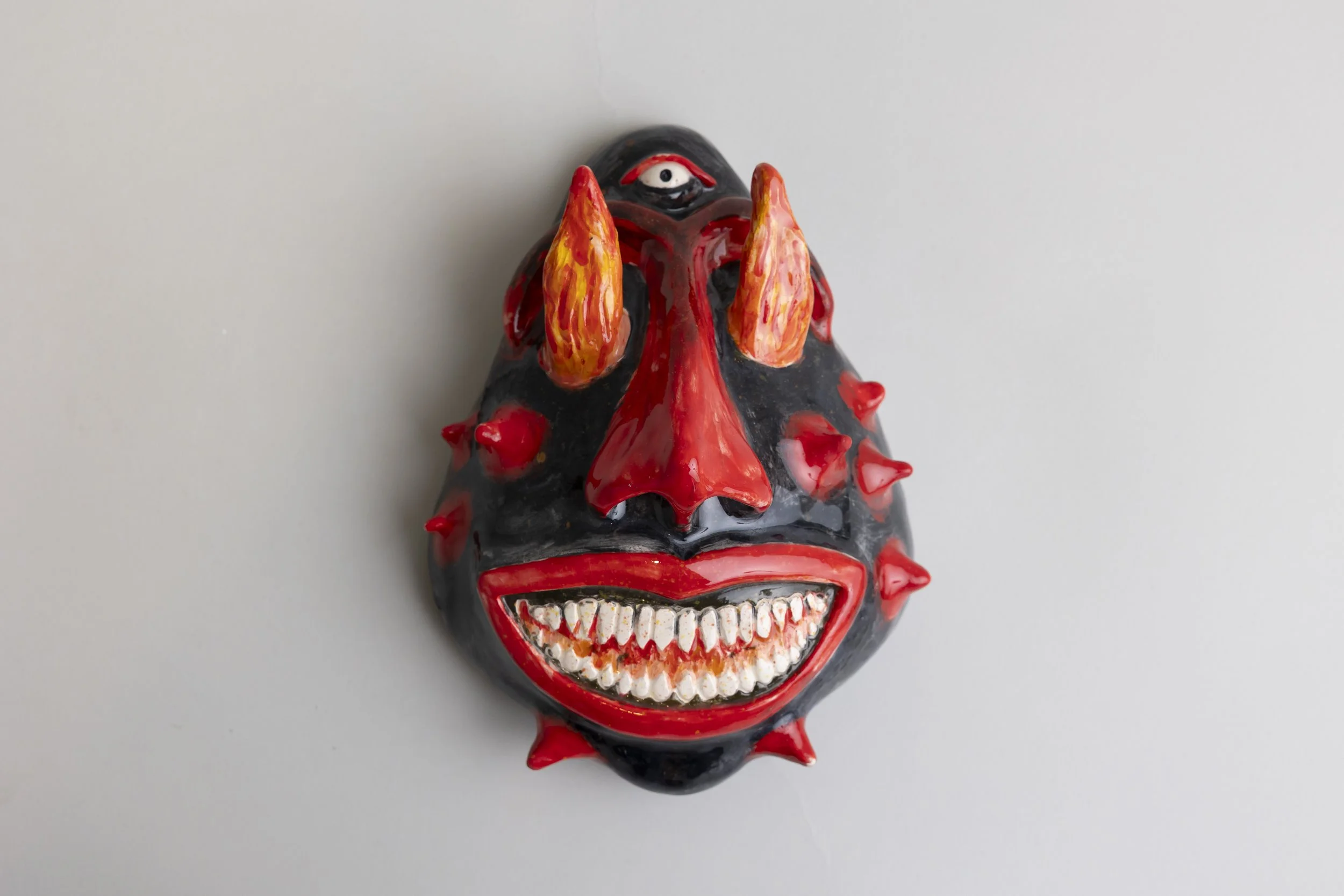 A colorful ceramic or painted sculpture of a clown face with exaggerated features, including red, yellow, and black colors, sharp teeth, and spiky details, against a plain background.