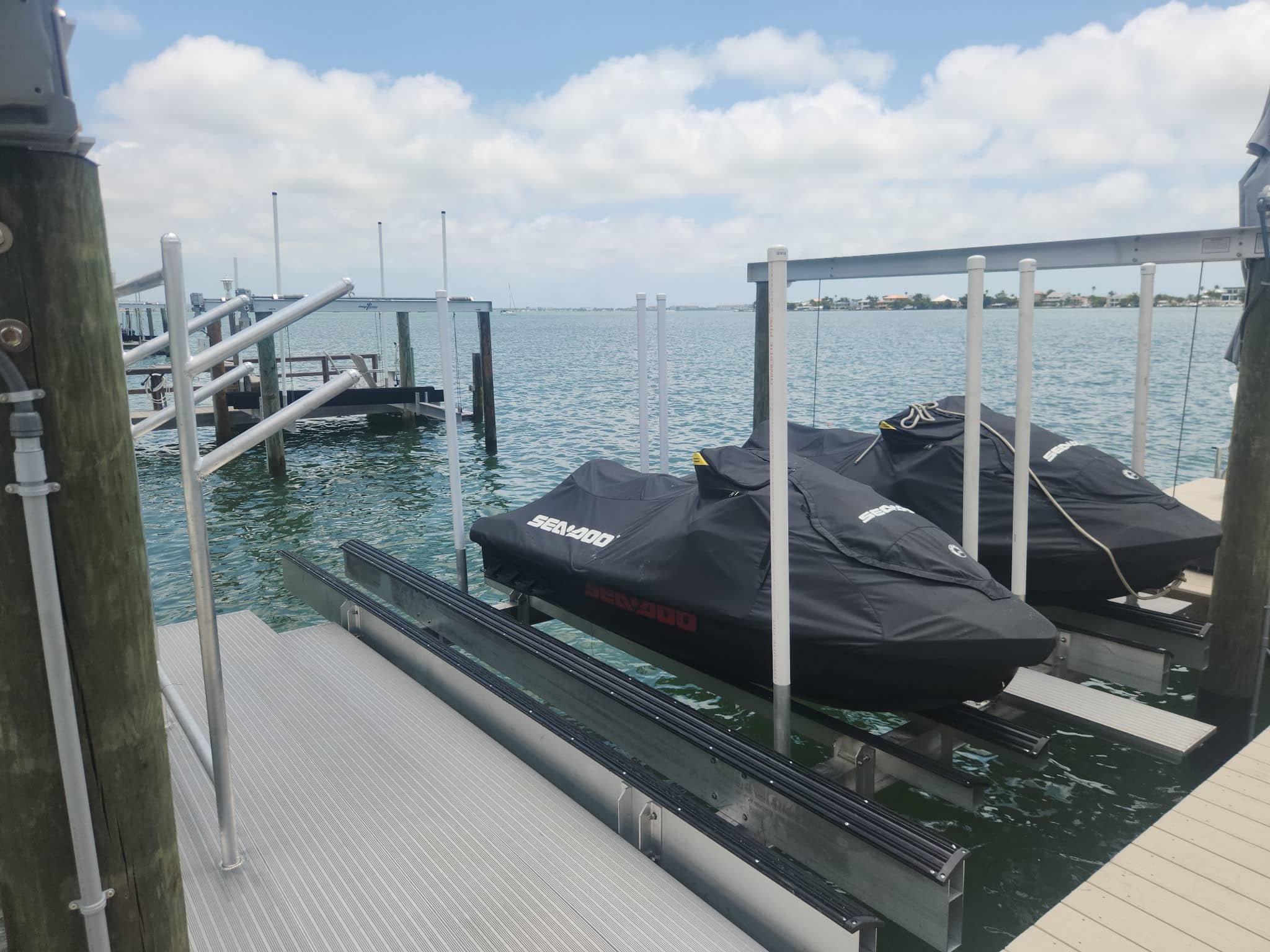 covered jet skis on a boat lift next to a dock with a platform in front of the ocean and a blue sky