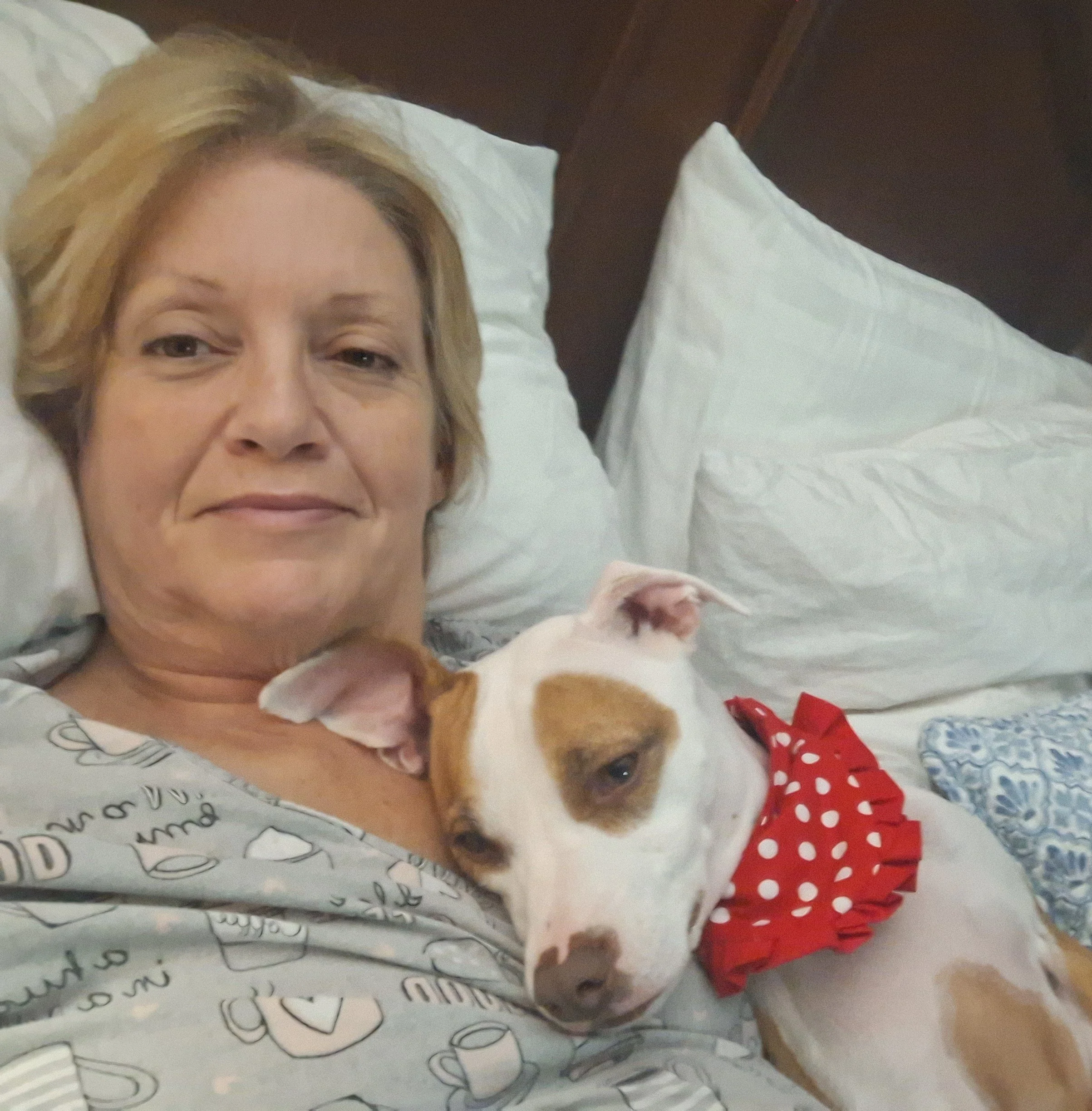 A woman lying in bed smiling, with a dog cuddled up bedside her, close-up of her face and the dog.