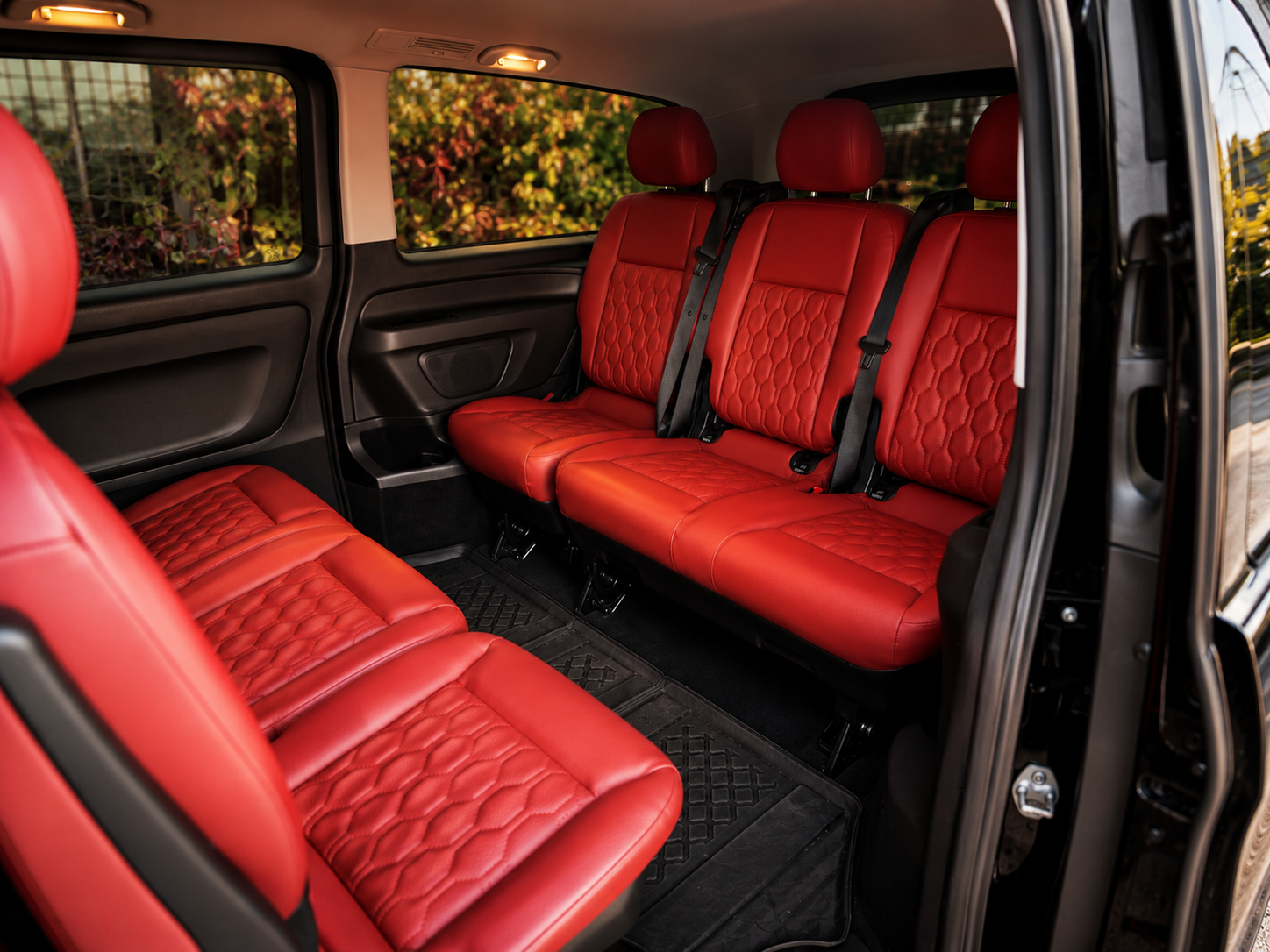 Interior of a vehicle with red leather seats and black seat belts, seen from an open door, with trees and a fence outside the windows.