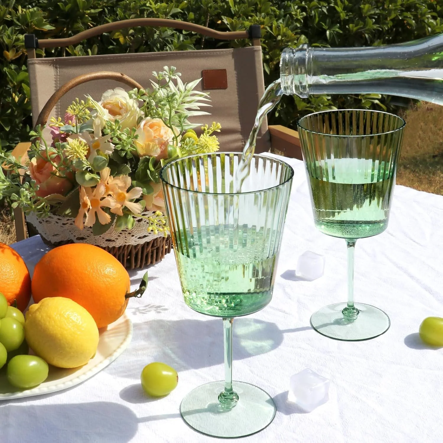 A table with a white tablecloth set outdoors, featuring two green glasses of water being filled from a pitcher, a bowl of various citrus fruits and green grapes, a tray with a bouquet of white and pink flowers, and two oranges in the foreground.