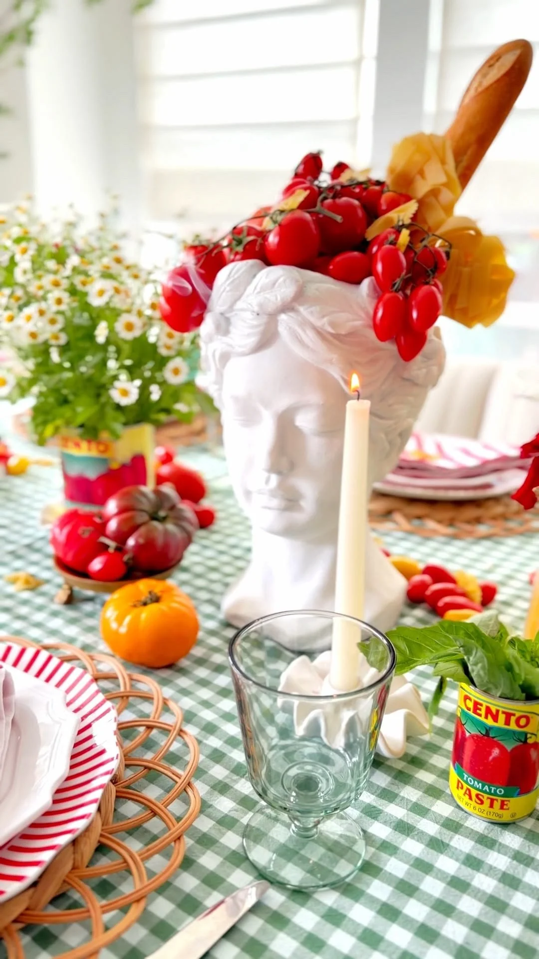 A table setting with a white bust flower vase topped with cherry tomatoes and yellow skewers, a candle, a glass of water, a can of tomato paste, a basket with a striped cloth, and a bunch of tomatoes on a green checkered tablecloth, with daisies in the background.