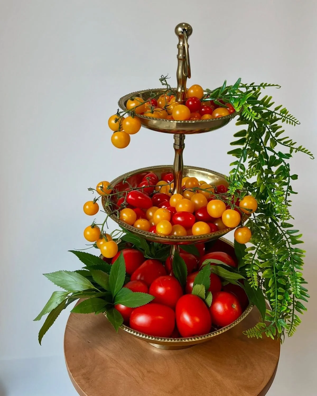 Three-tiered metal fruit stand filled with cherry tomatoes of various colors and green foliage, set on a wooden table with a plain white wall background.