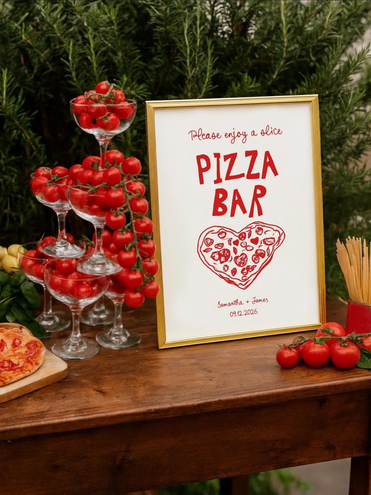 A table with cherry tomatoes in glass cups arranged in a pyramid, a framed sign for a pizza bar, and a small pizza slice on a cutting board, all outdoors with greenery in the background.