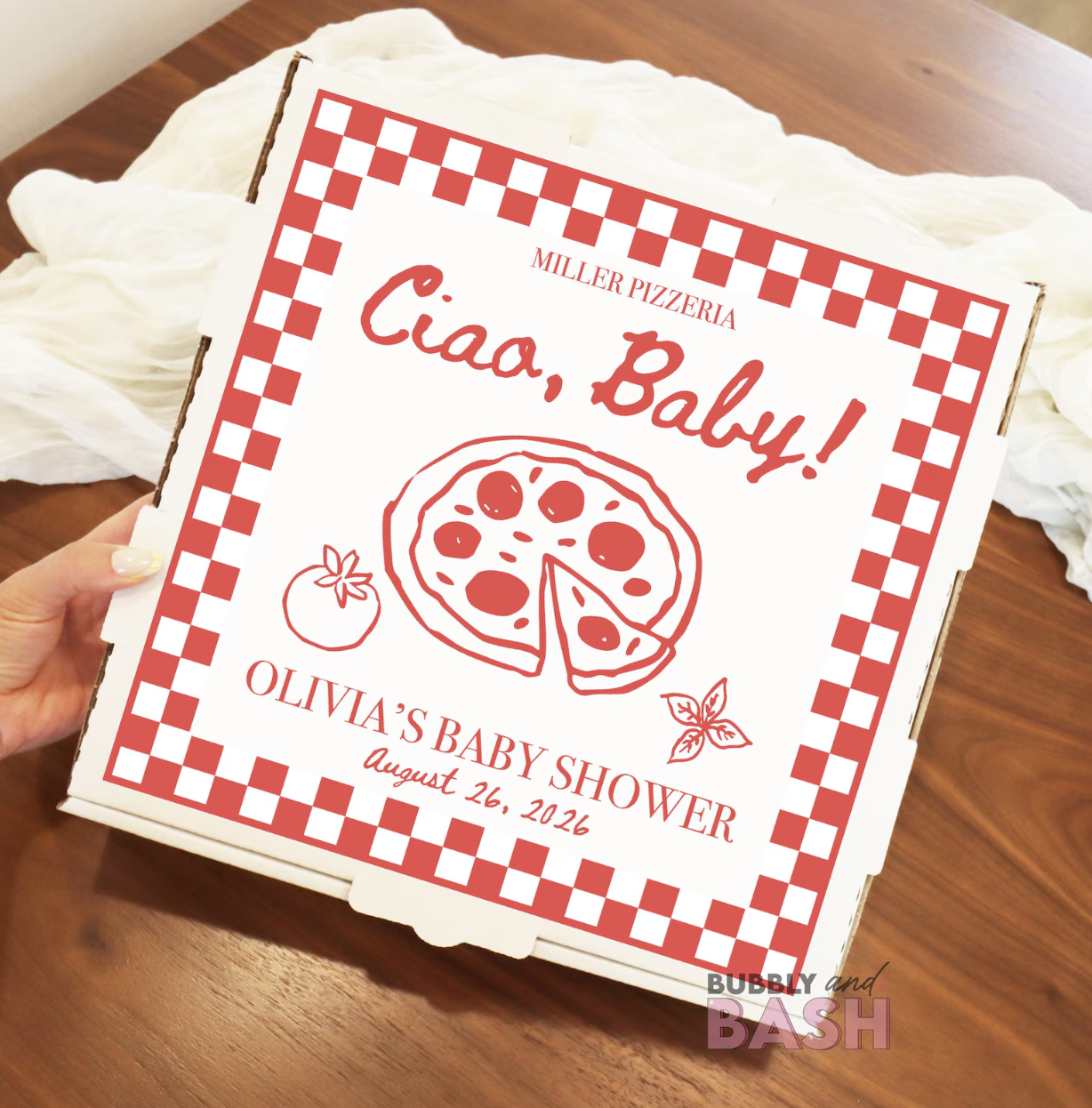 Pizza box with red checkered border, featuring text 'Ciao, Baby!', a drawing of a pizza, and details about a baby shower on August 26, 2026, with a tomato and basil illustration.
