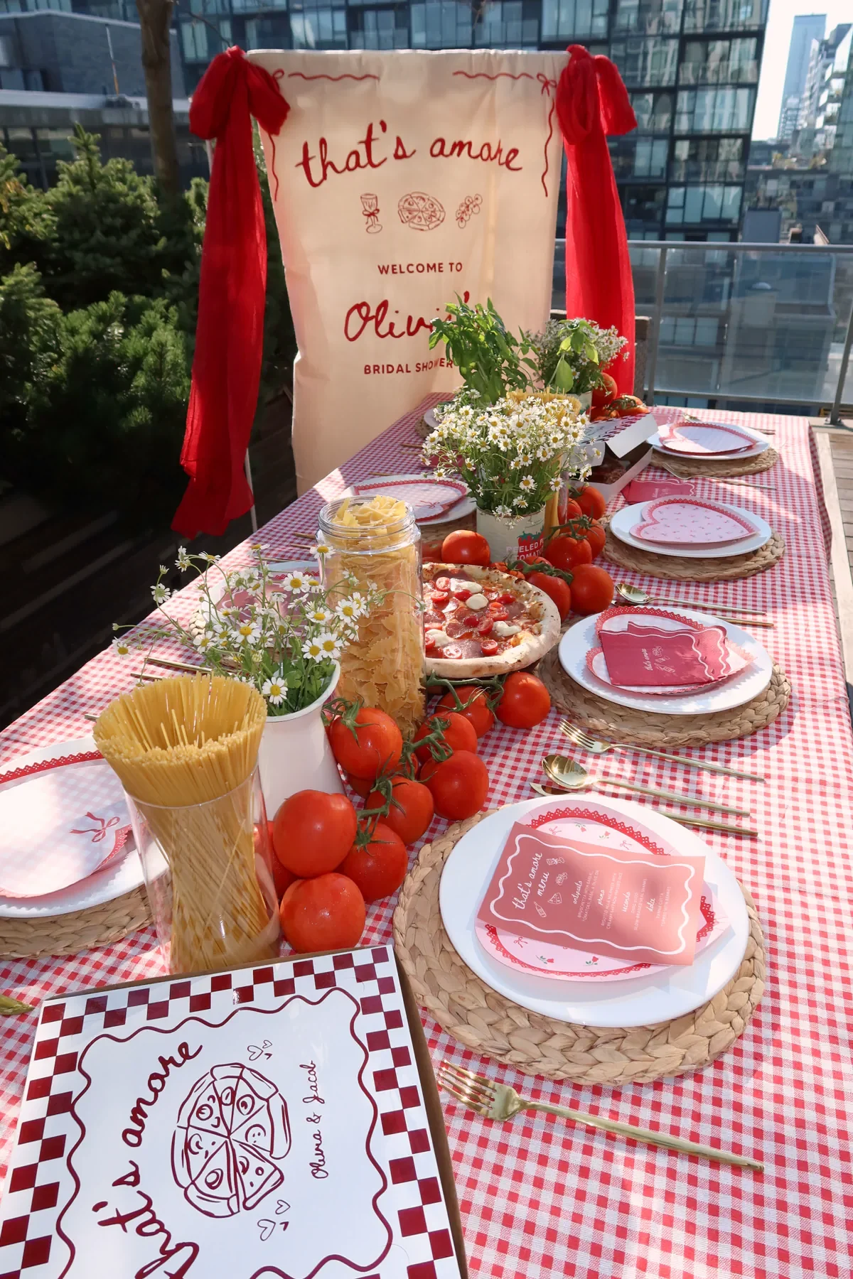 A tablescape decorated with a red and white checkered tablecloth, featuring a bouquet of daisies, fresh tomatoes, a pizza, and various tableware, with a decorative backdrop that says 'that's amore' welcoming Olivia for a bridal shower.