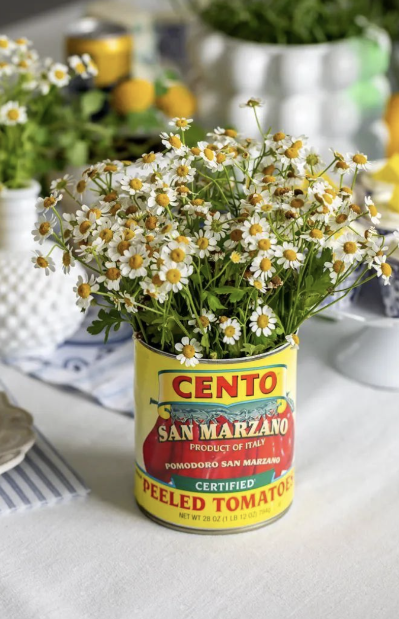 A bouquet of white daisies with yellow centers in a yellow can labeled 'Cento San Marzano Peeled Tomatoes'. The can is used as a vase, and there are other flowers and cans in the blurred background.