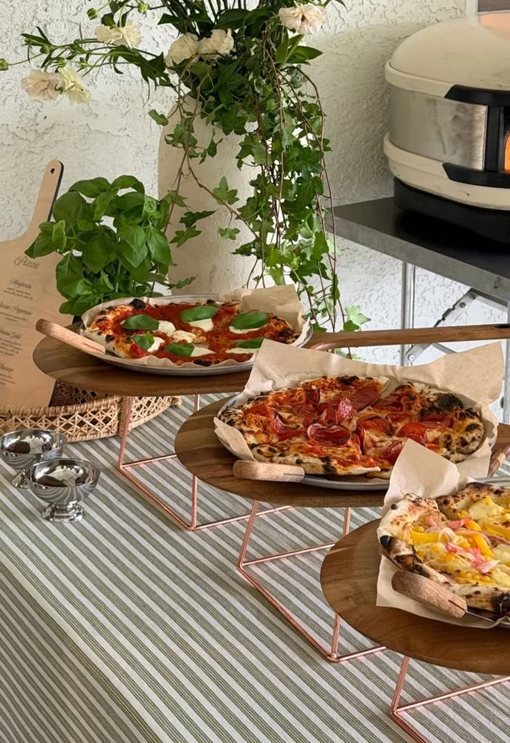 Three pizzas on wooden serving stands on a striped tablecloth, with a plant and pizza oven in the background.